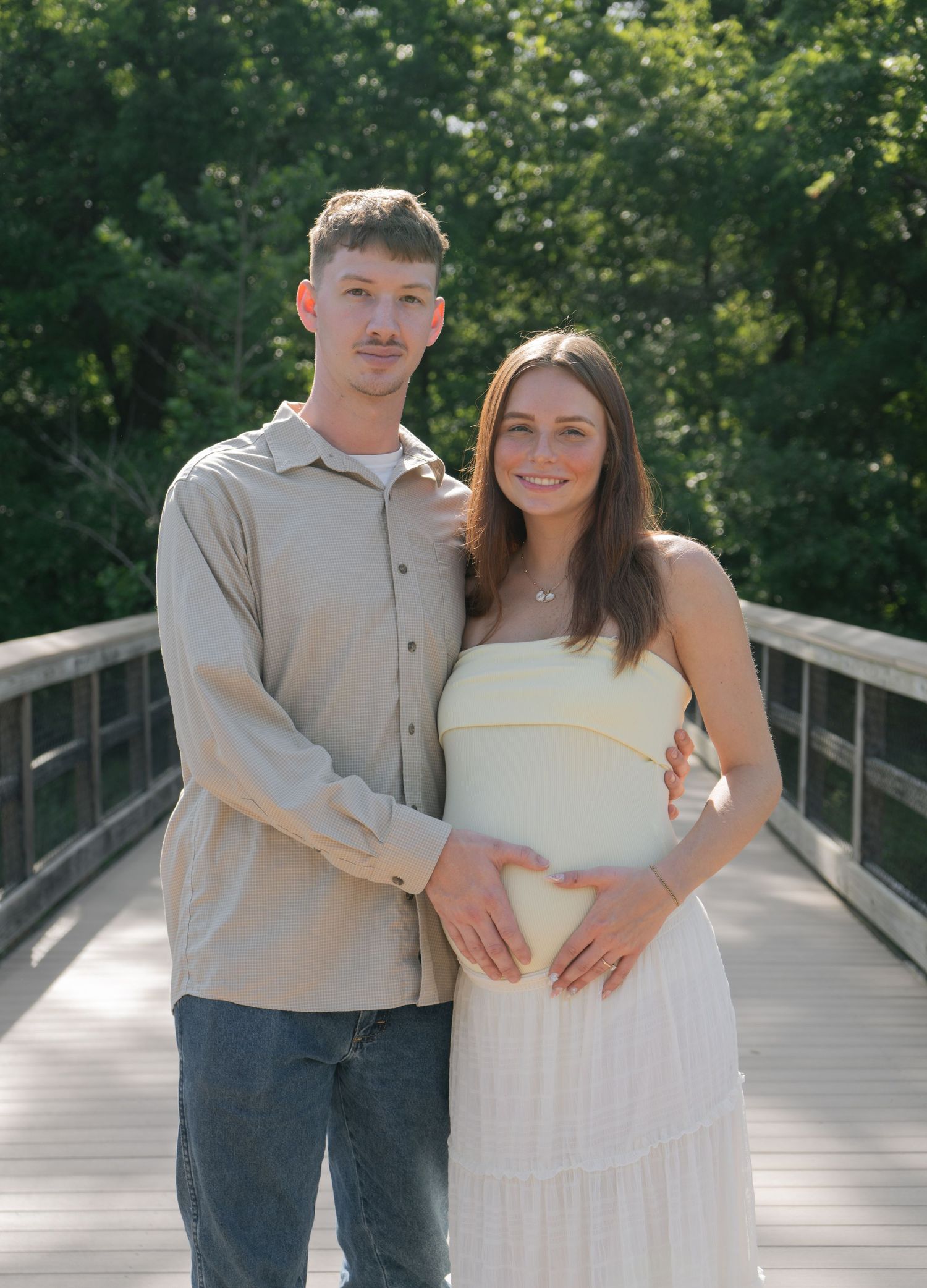 Couple posing on a wooden bridge outdoors, woman in white maternity dress, man in beige shirt, green trees background.