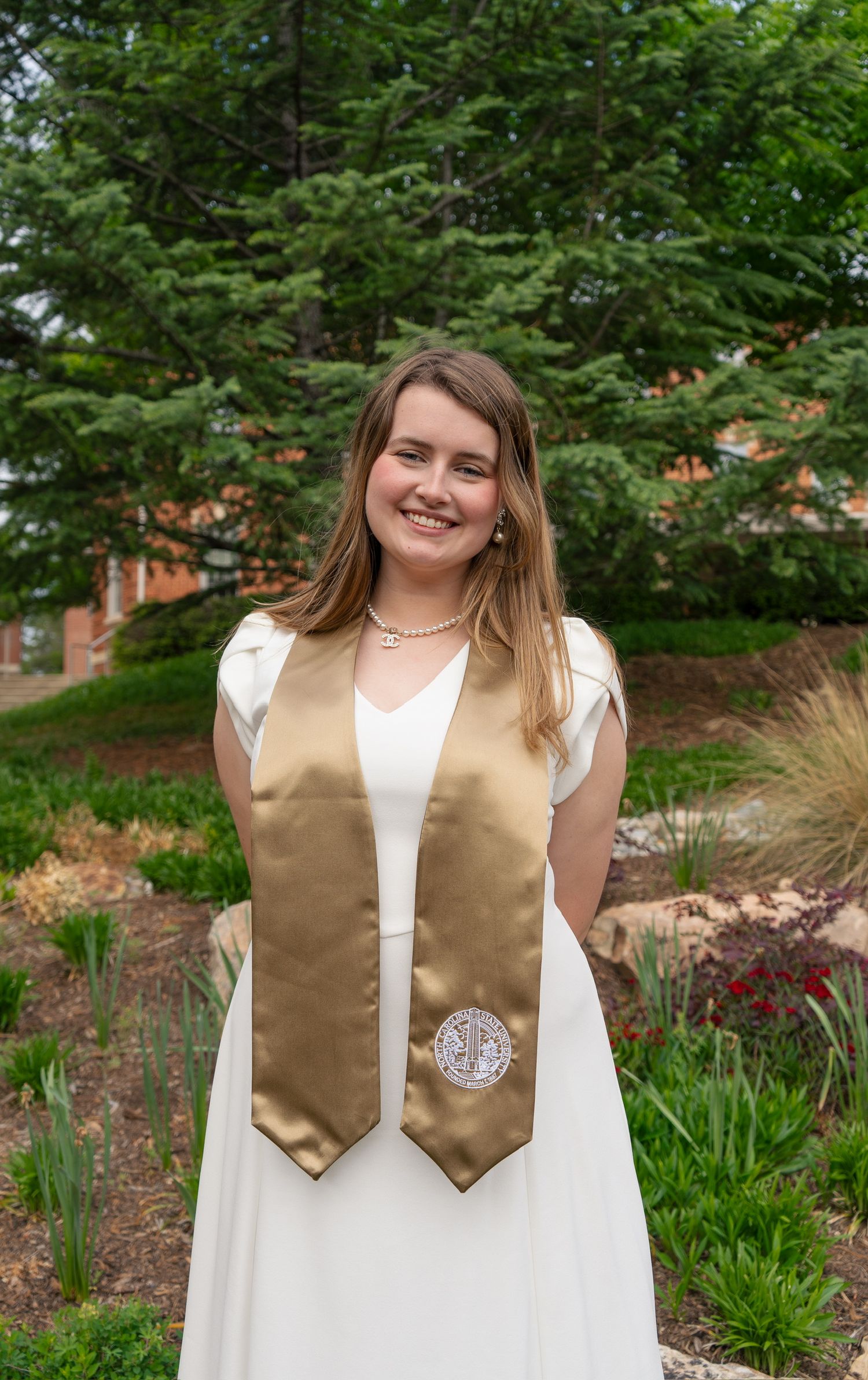 Smiling graduate in white dress with gold university stole poses in front of green campus garden.