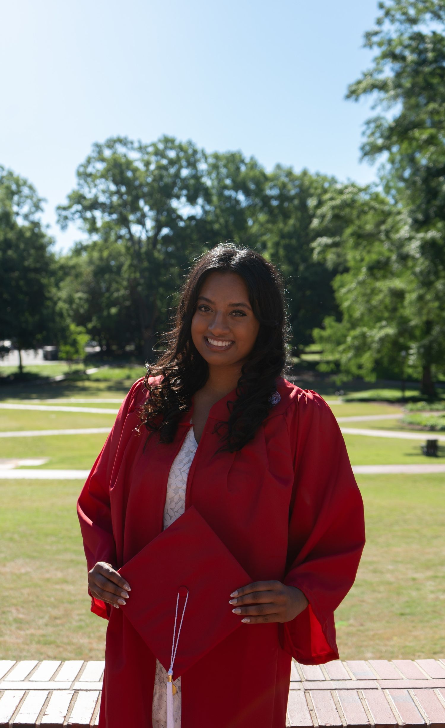 Smiling young woman in red graduation gown holds cap while standing outdoors on sunny day with green trees.