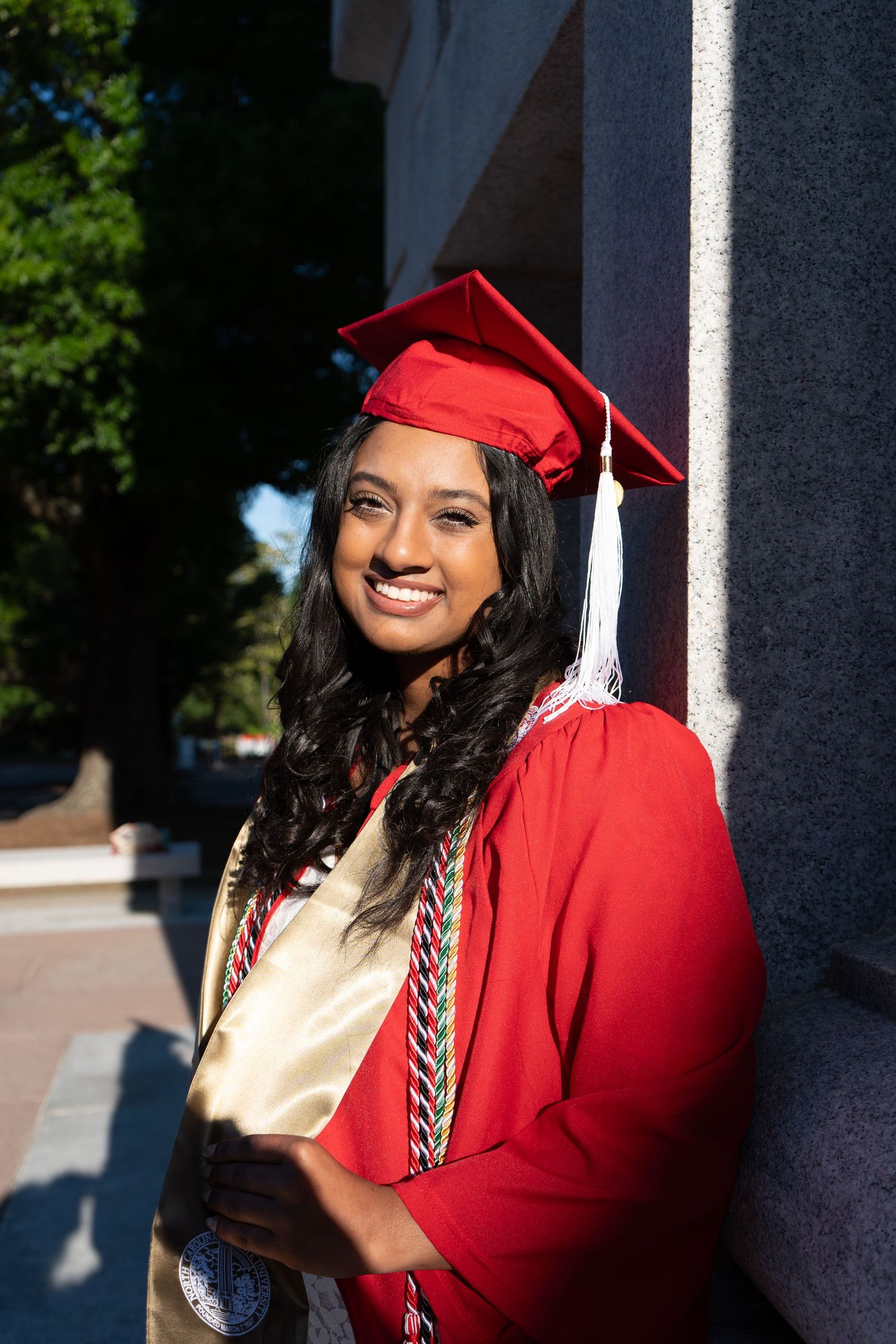 Smiling graduate in red cap and gown leaning against a pillar outdoors on a sunny day.