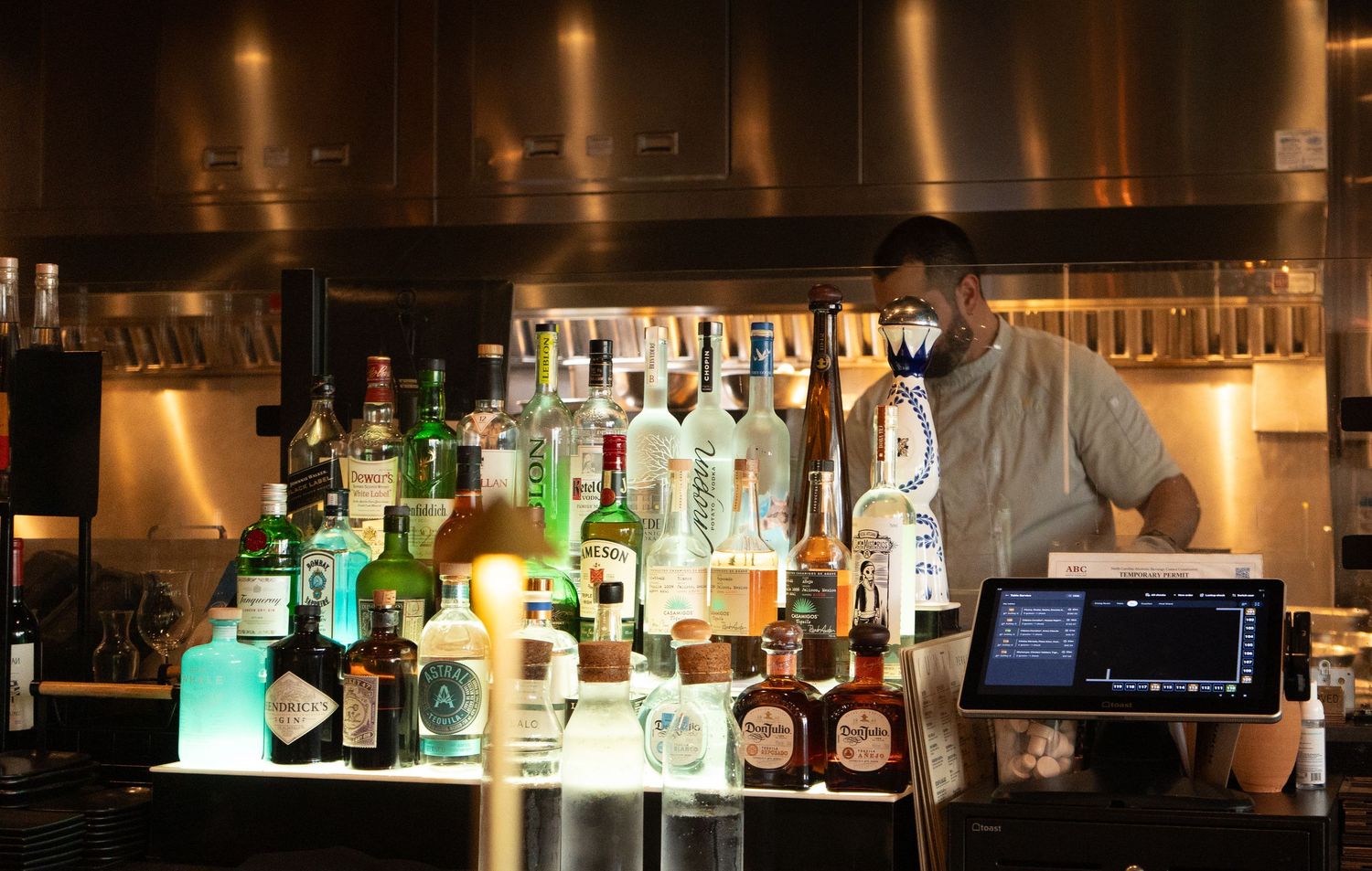 Professional branding photographer captures bartender standing behind a well-stocked bar with colorful liquor bottles and a POS system.
