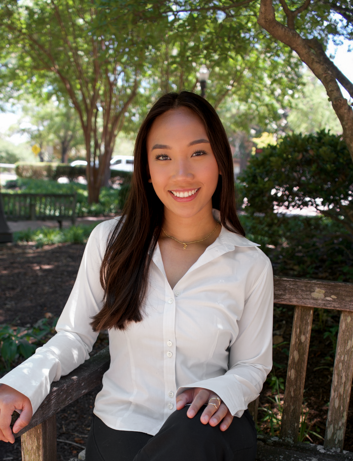 Smiling young woman in white blouse sits on a bench outdoors surrounded by lush green trees.