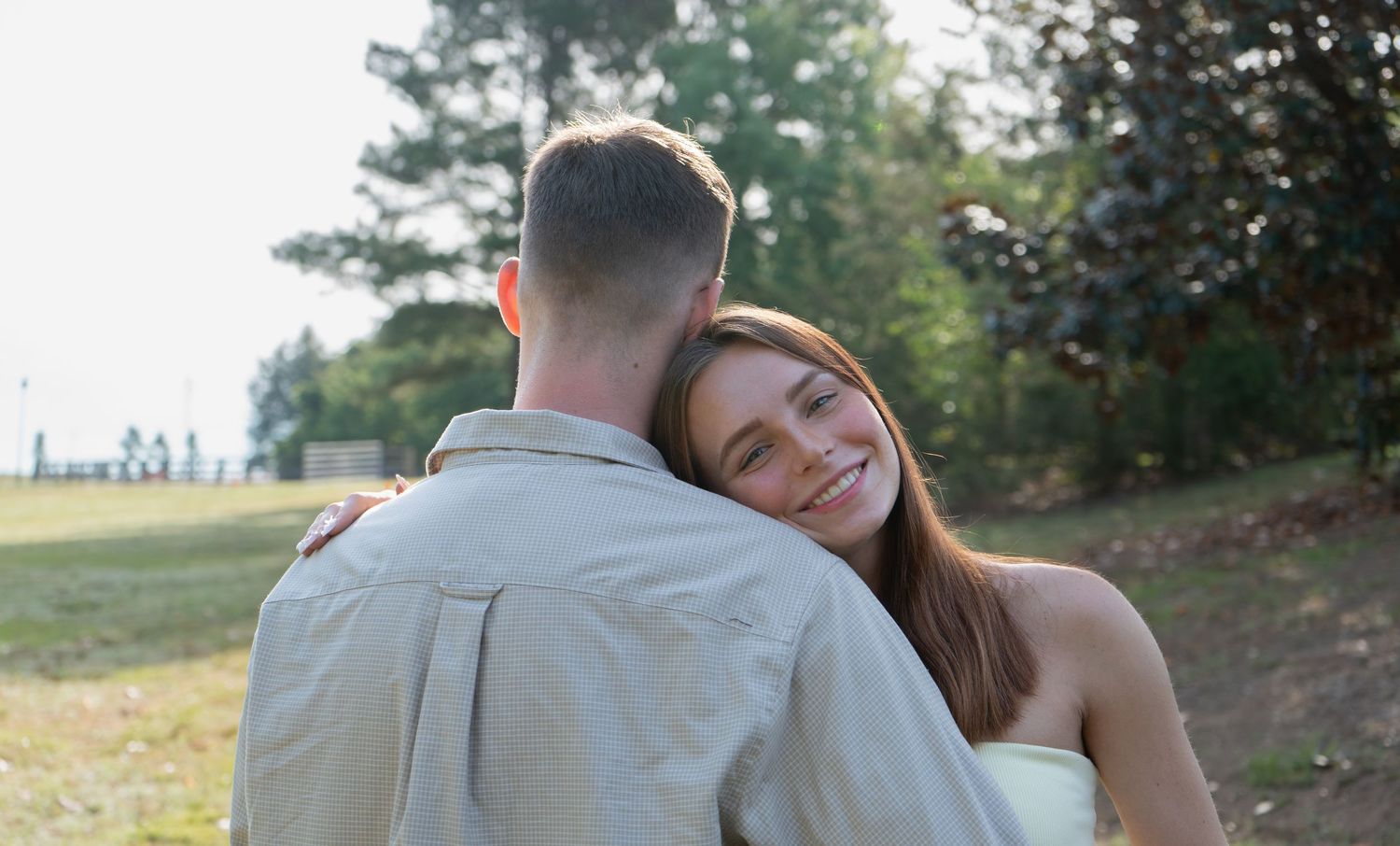 A smiling young woman hugs a man outdoors in a park setting with green trees in the background.