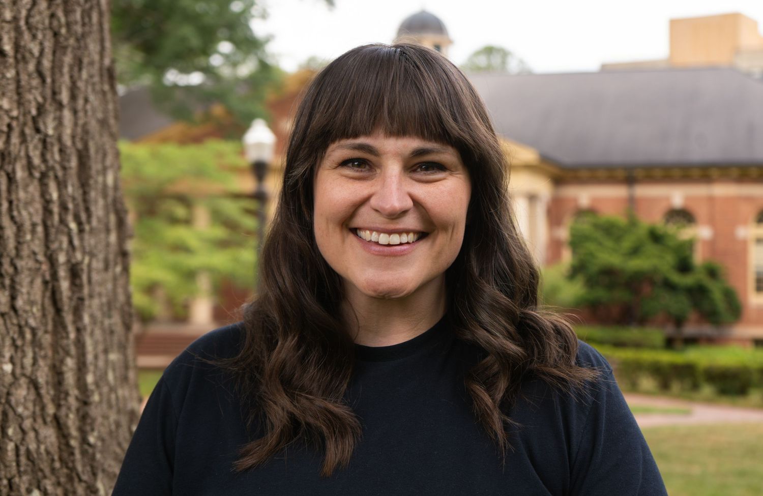 Smiling woman with dark hair and bangs standing outdoors near a tree on NC State campus.