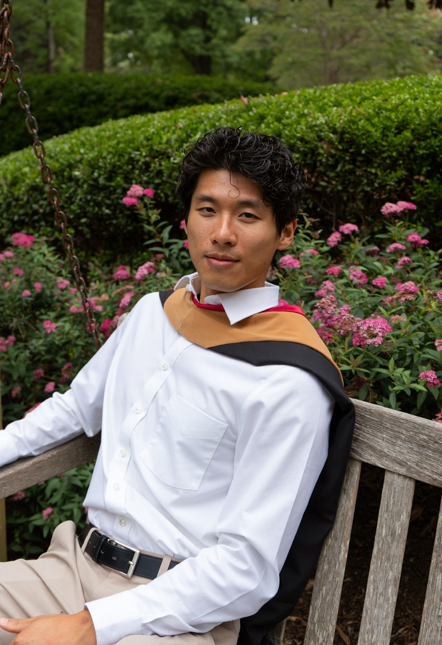 Young man in white dress shirt with graduation hood sits on bench surrounded by blooming flowers.