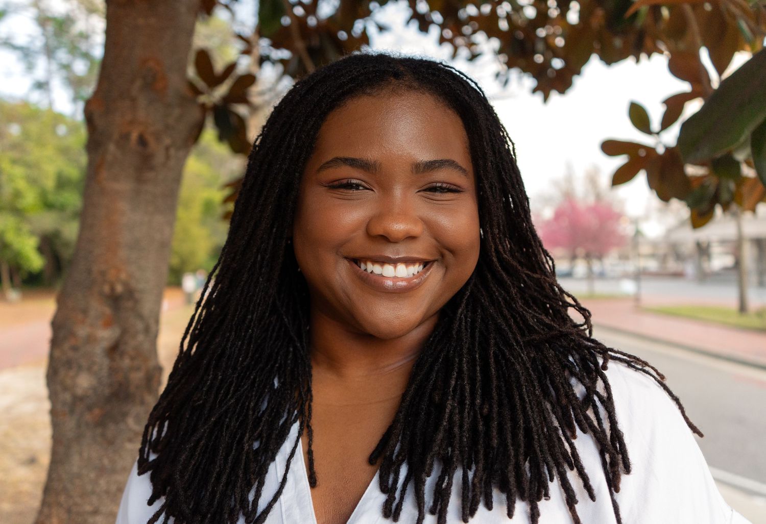 Happy young woman with long locs smiles broadly outdoors near colorful spring trees.
