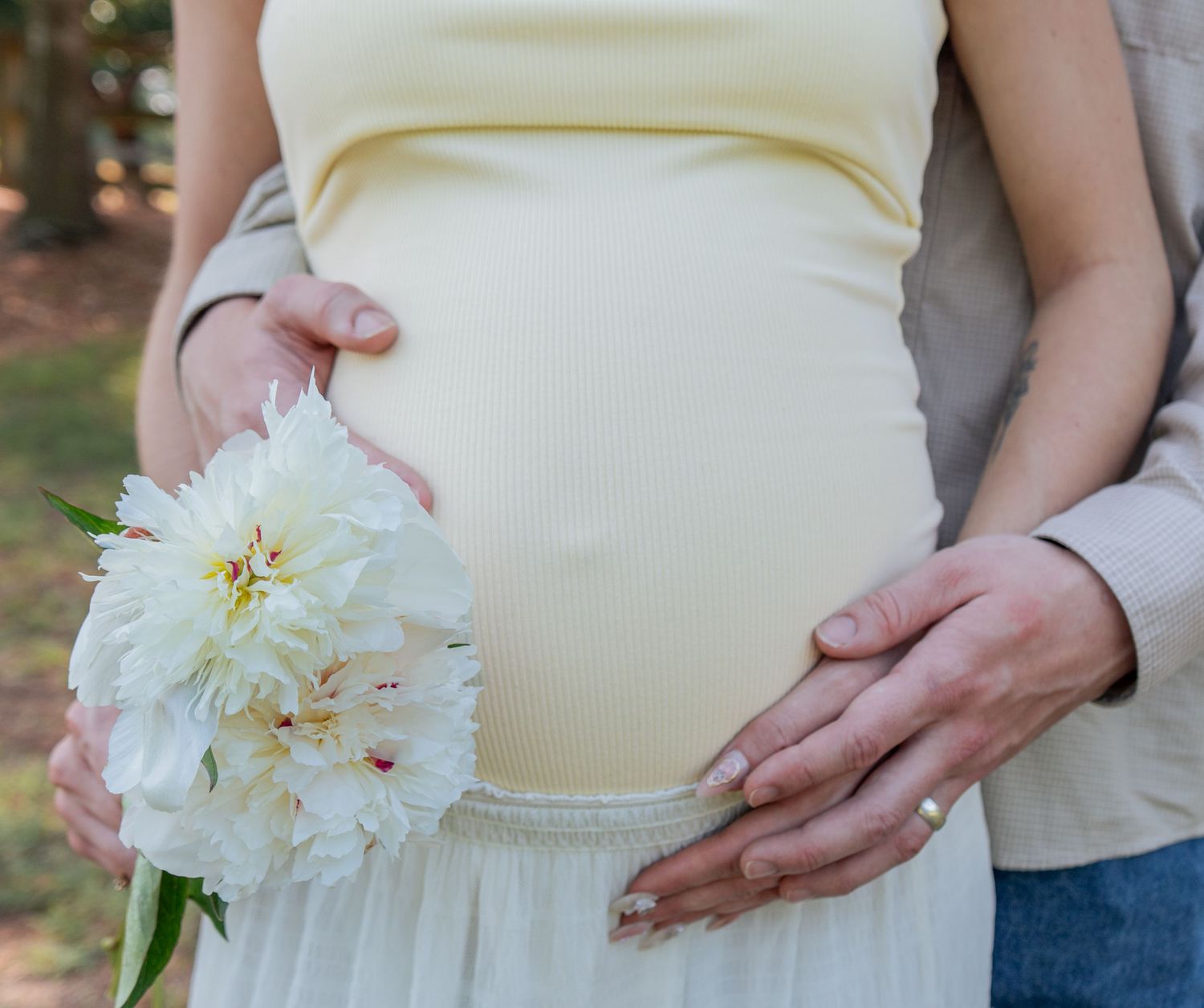 Pregnant woman holding white peony flowers with partner's hands on baby bump outdoors.