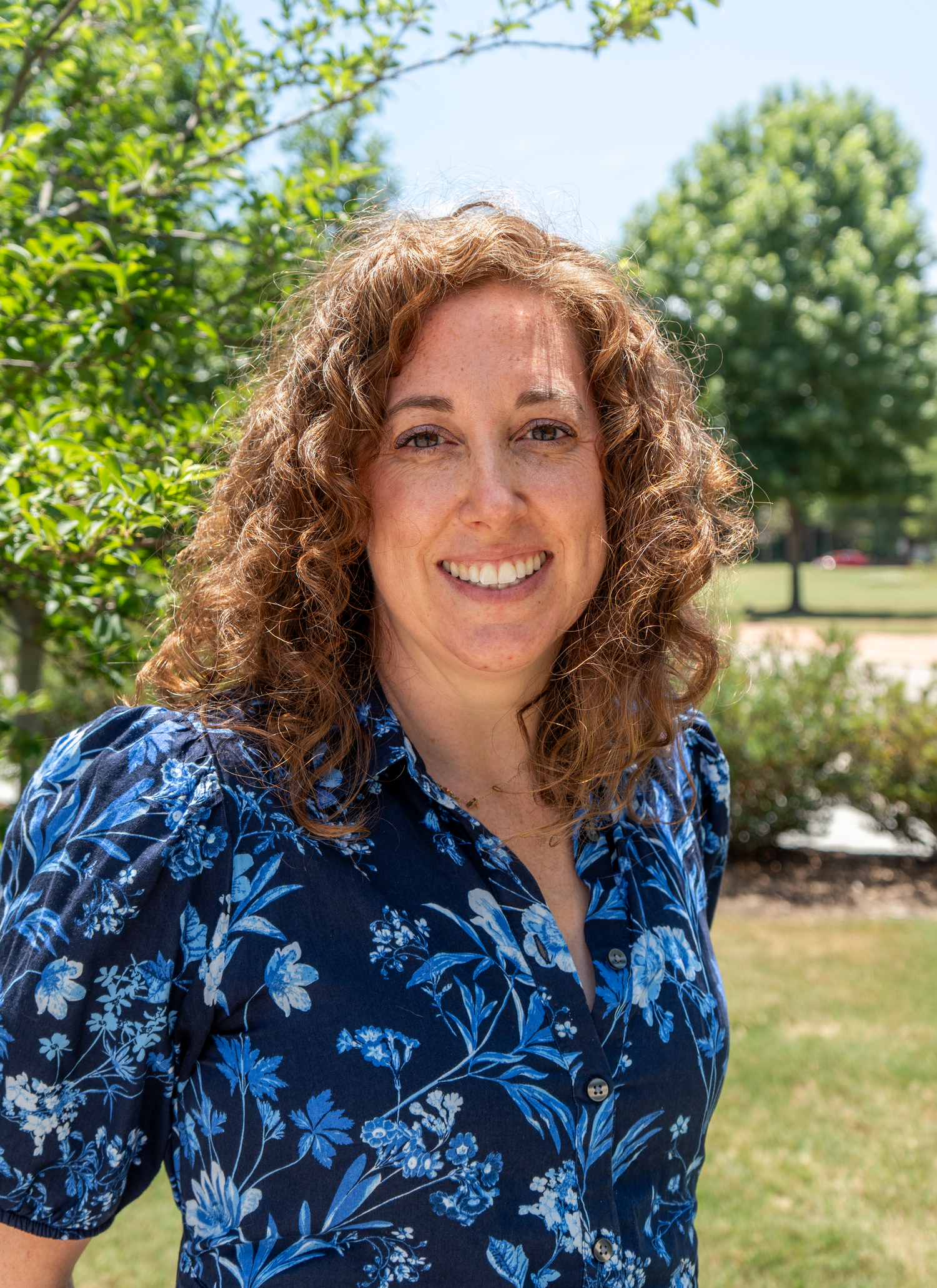 Woman with curly hair smiling brightly outdoors wearing blue floral blouse with green trees behind her.