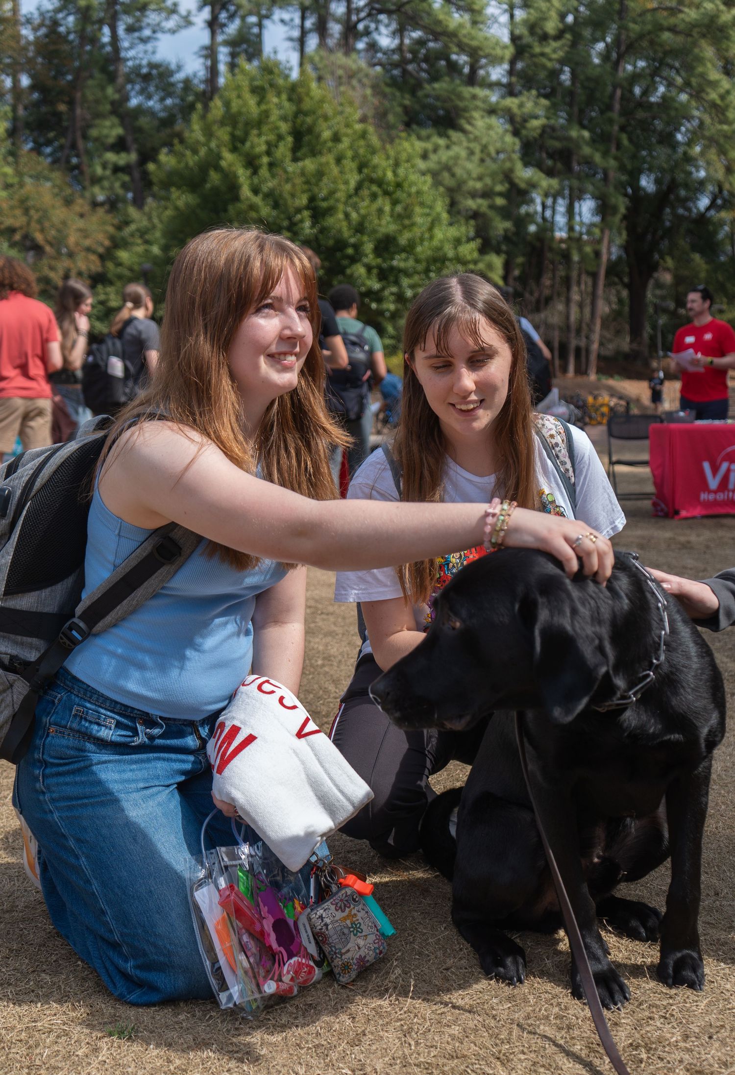 Two smiling female students kneel to pet a black Labrador dog at an outdoor campus event.