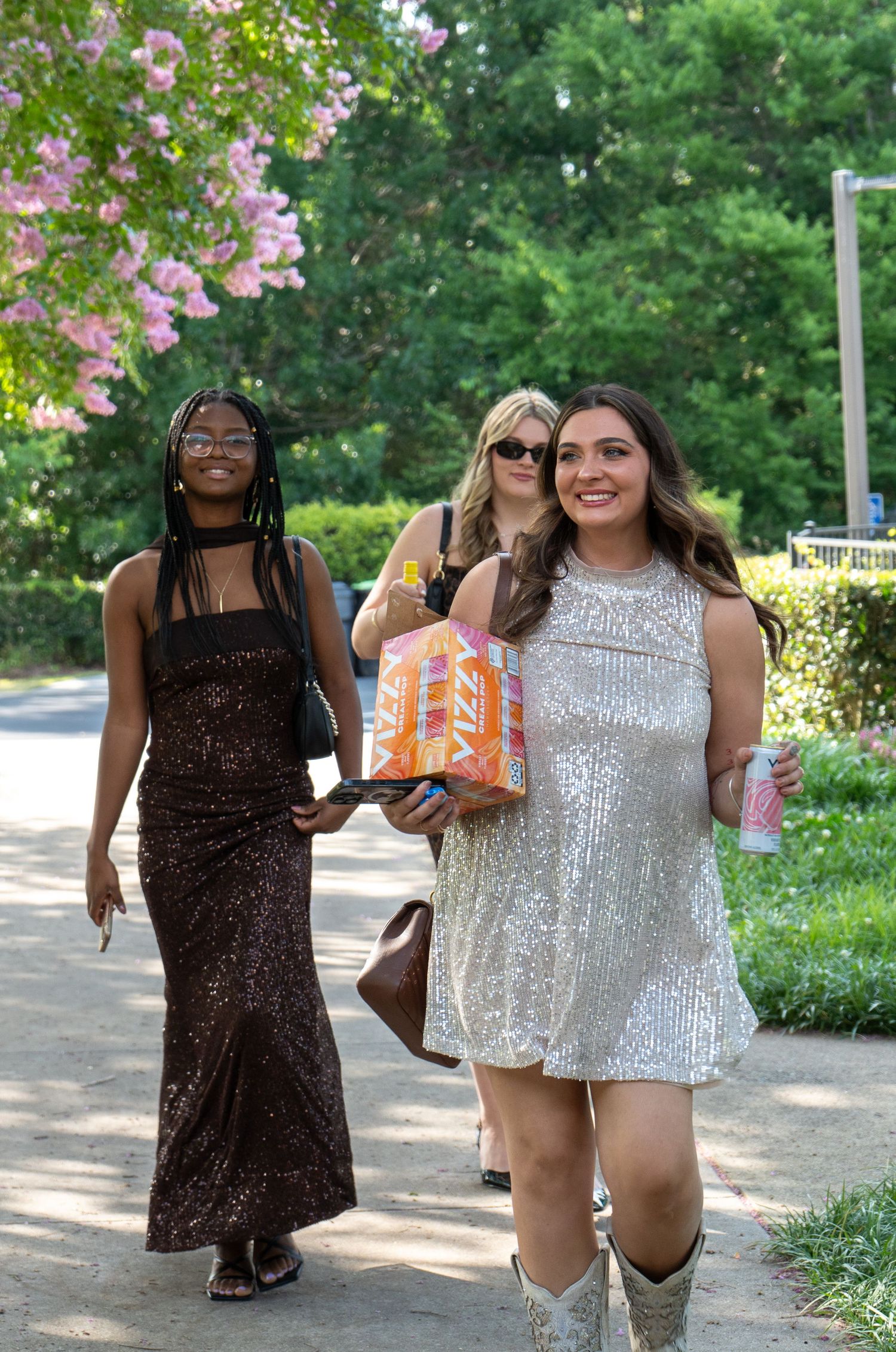 Three women in glamorous sequin dresses walk along a sunny outdoor path.