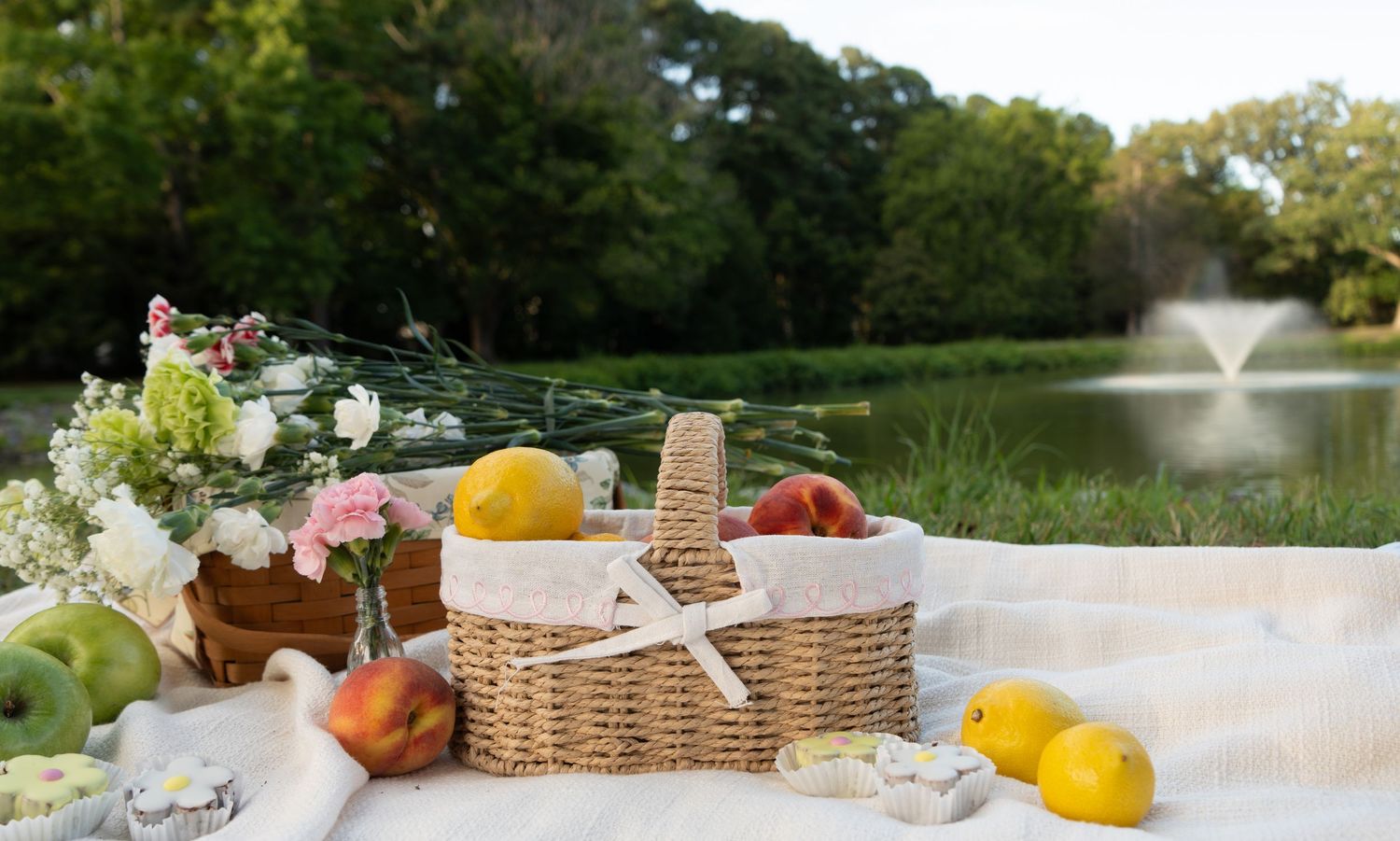 Picnic blanket with wicker basket, fresh peaches, lemons, and flowers by a serene pond with a fountain.