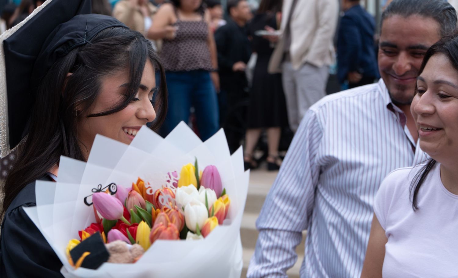 Graduate in cap and gown receiving colorful flower bouquet from proud family members outdoors.