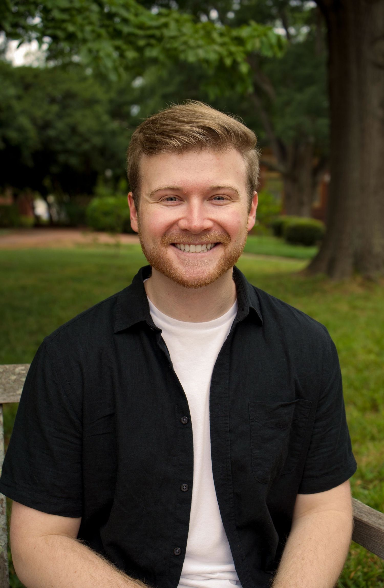 Young man with red beard smiling while seated on a bench in a lush green park setting.