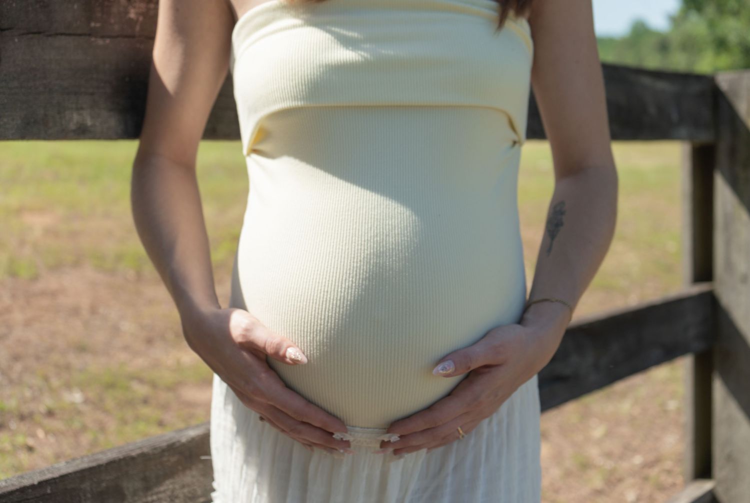 Pregnant woman in white dress cradling baby bump outdoors near wooden fence on sunny day.