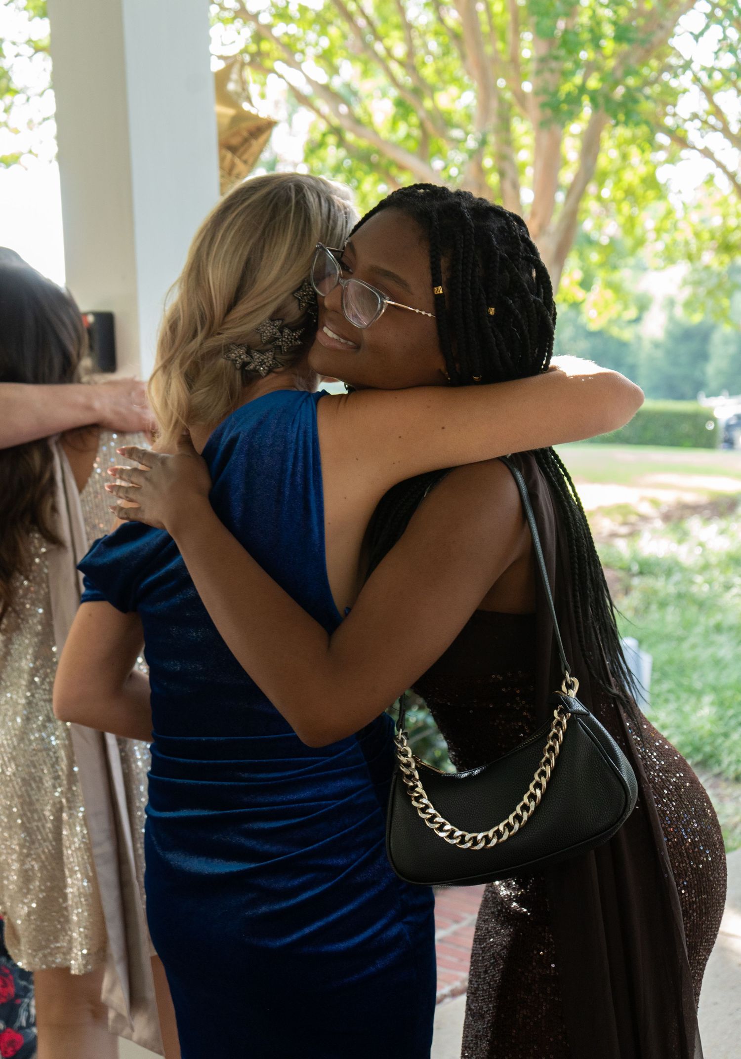 Two women in elegant dresses share a warm hug at an outdoor event.