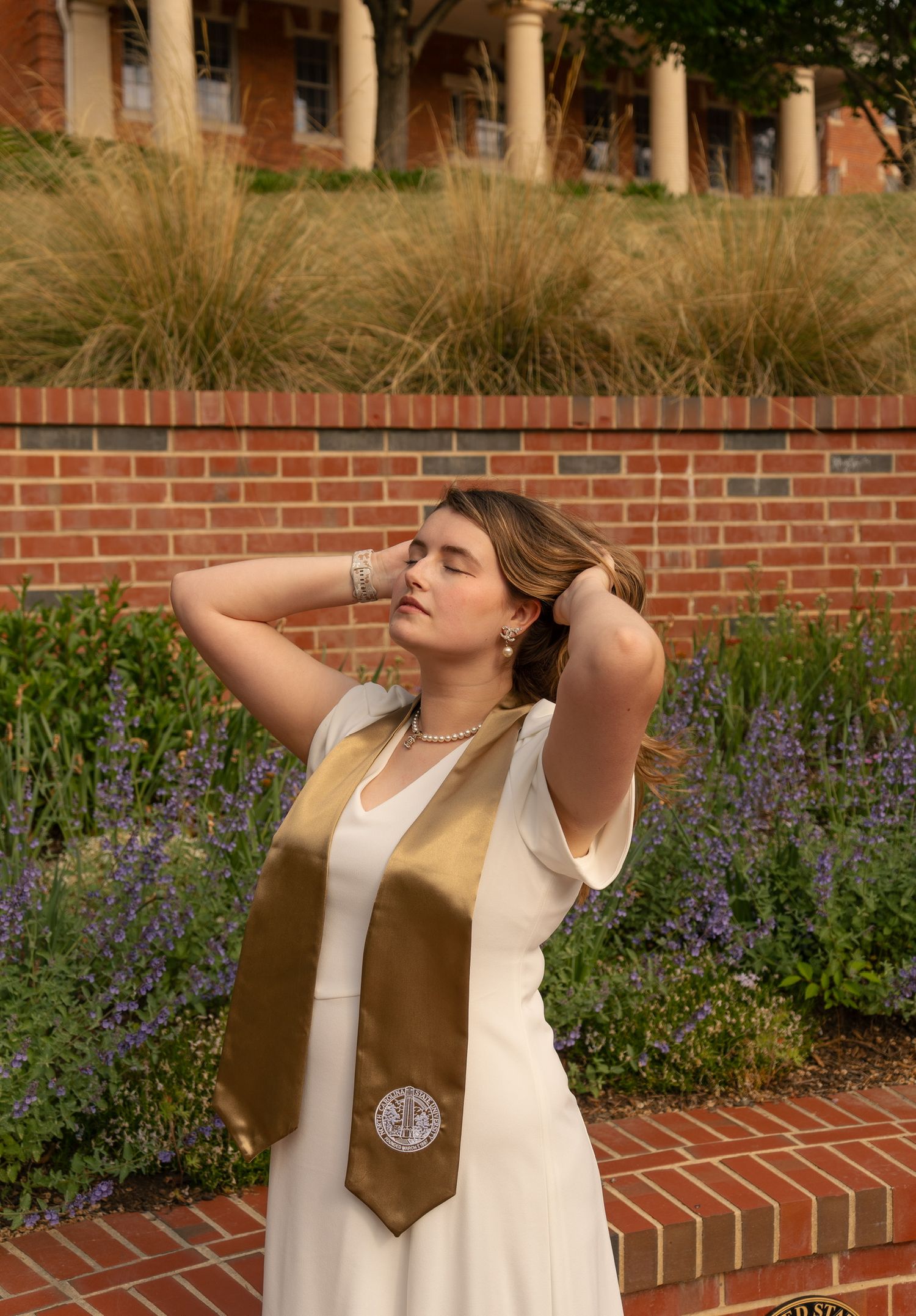 Graduate in gold stole and white dress poses with eyes closed near brick wall and garden.