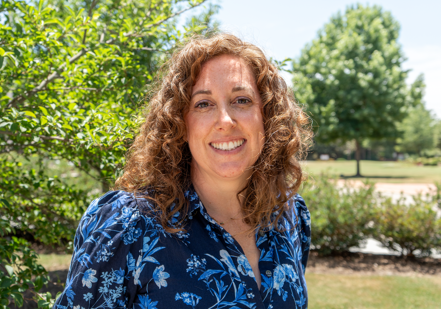 Smiling woman with curly auburn hair wearing blue floral blouse standing outdoors among green trees.