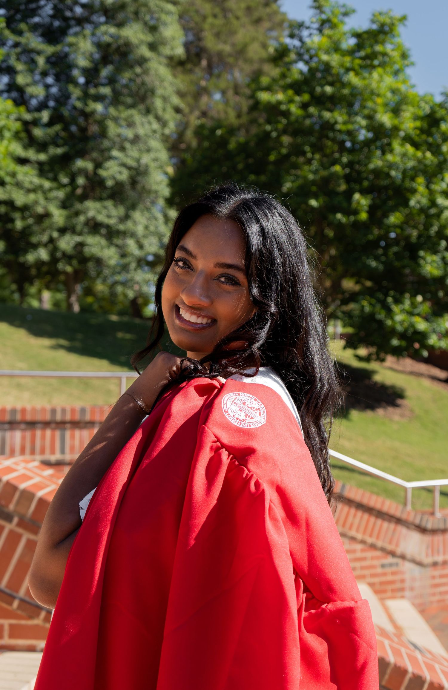 Young Indian woman smiling over her shoulder wearing a red graduation gown outdoors.
