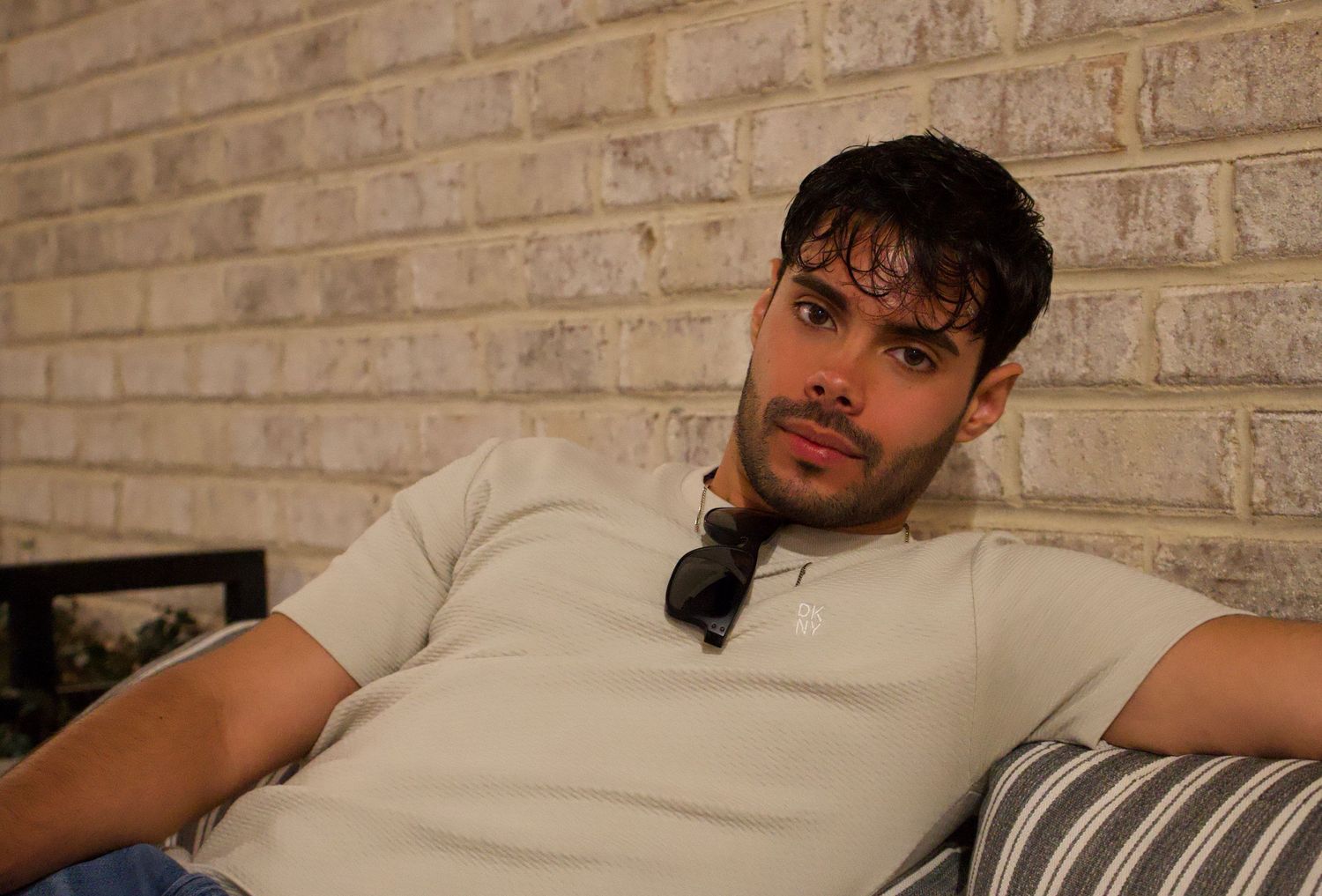 A young man with dark hair relaxes on a striped couch against a brick wall wearing a light gray shirt.