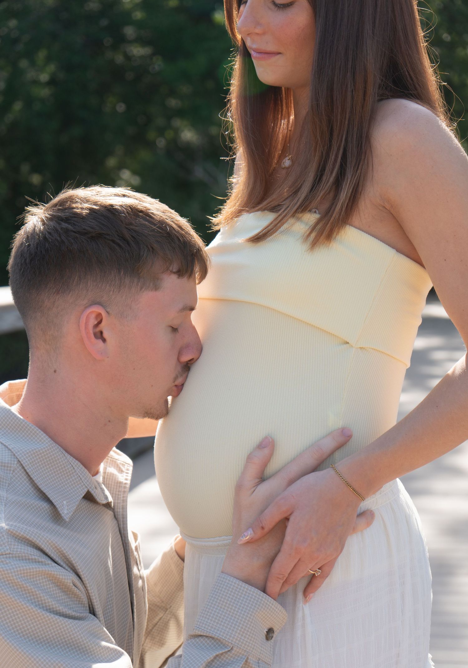 A man tenderly kisses a pregnant woman's baby bump during an outdoor maternity photo shoot.