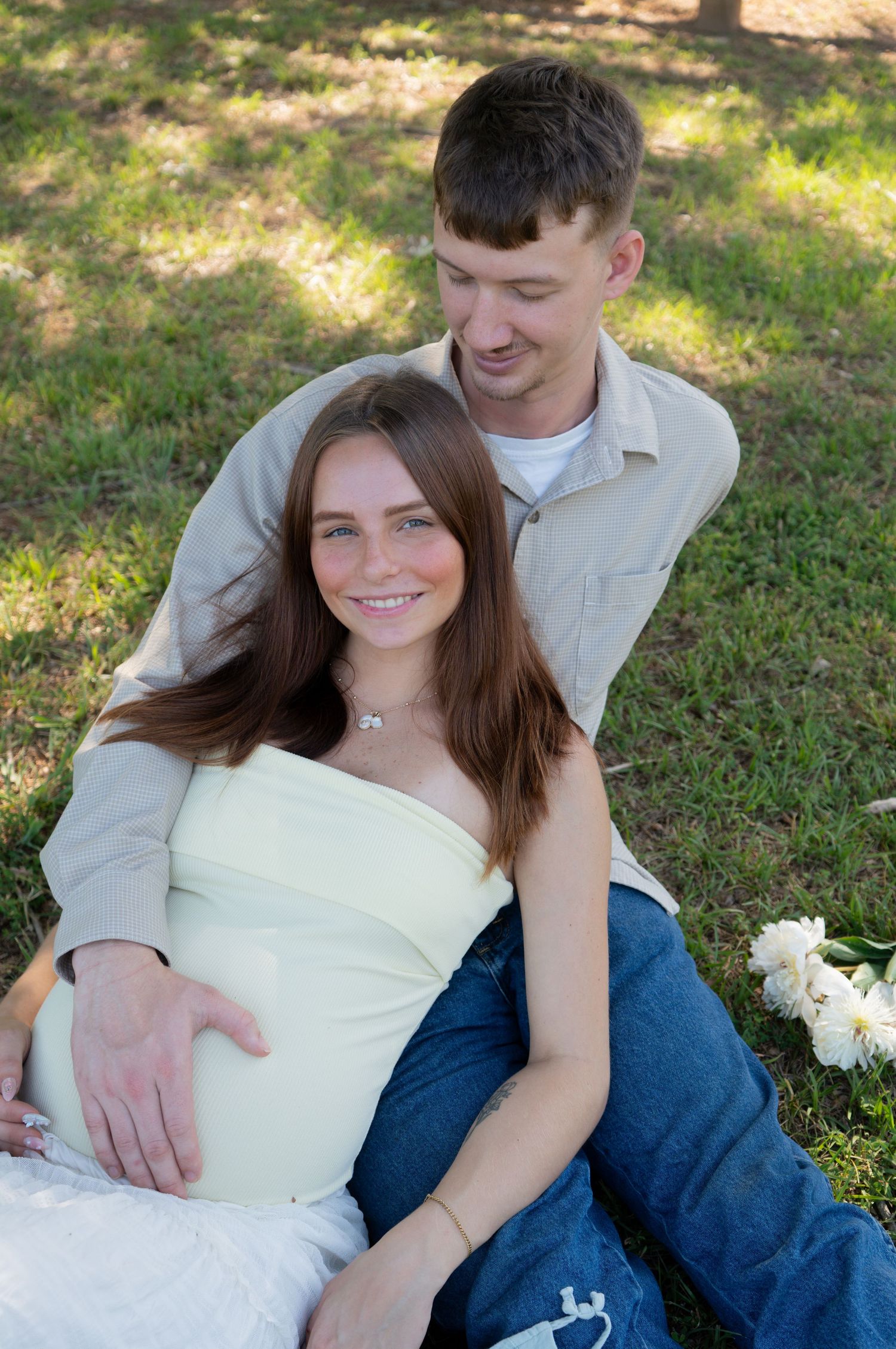 Pregnant woman in white top sits with partner on grass outdoors during a maternity photo session.