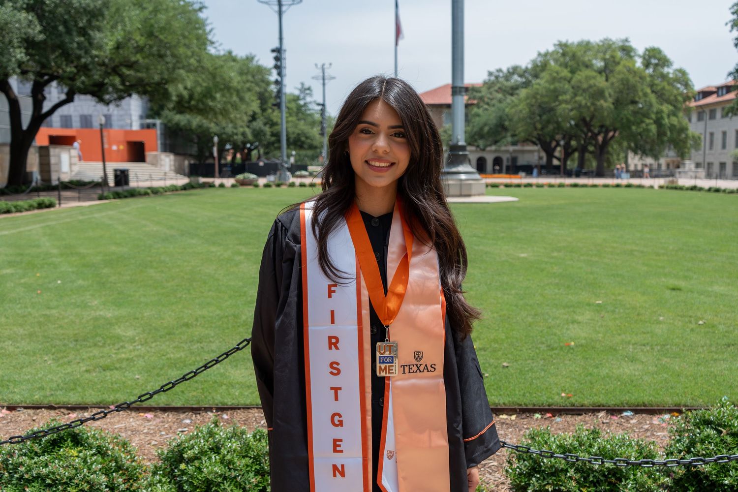 Female graduate in black gown with orange First Gen sash posing on university campus lawn.