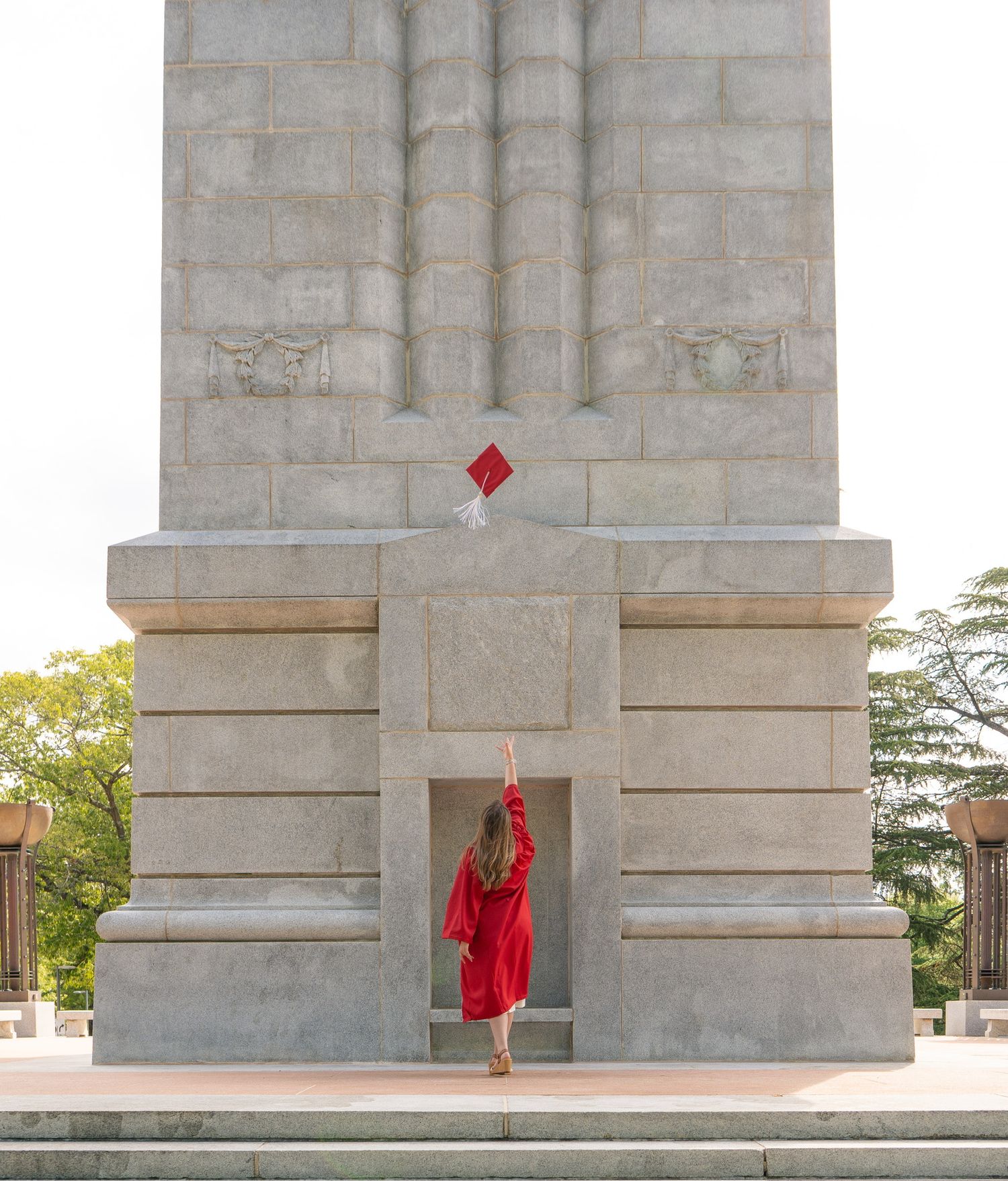 Graduate in red gown tosses cap at base of large stone monument or memorial tower outdoors.
