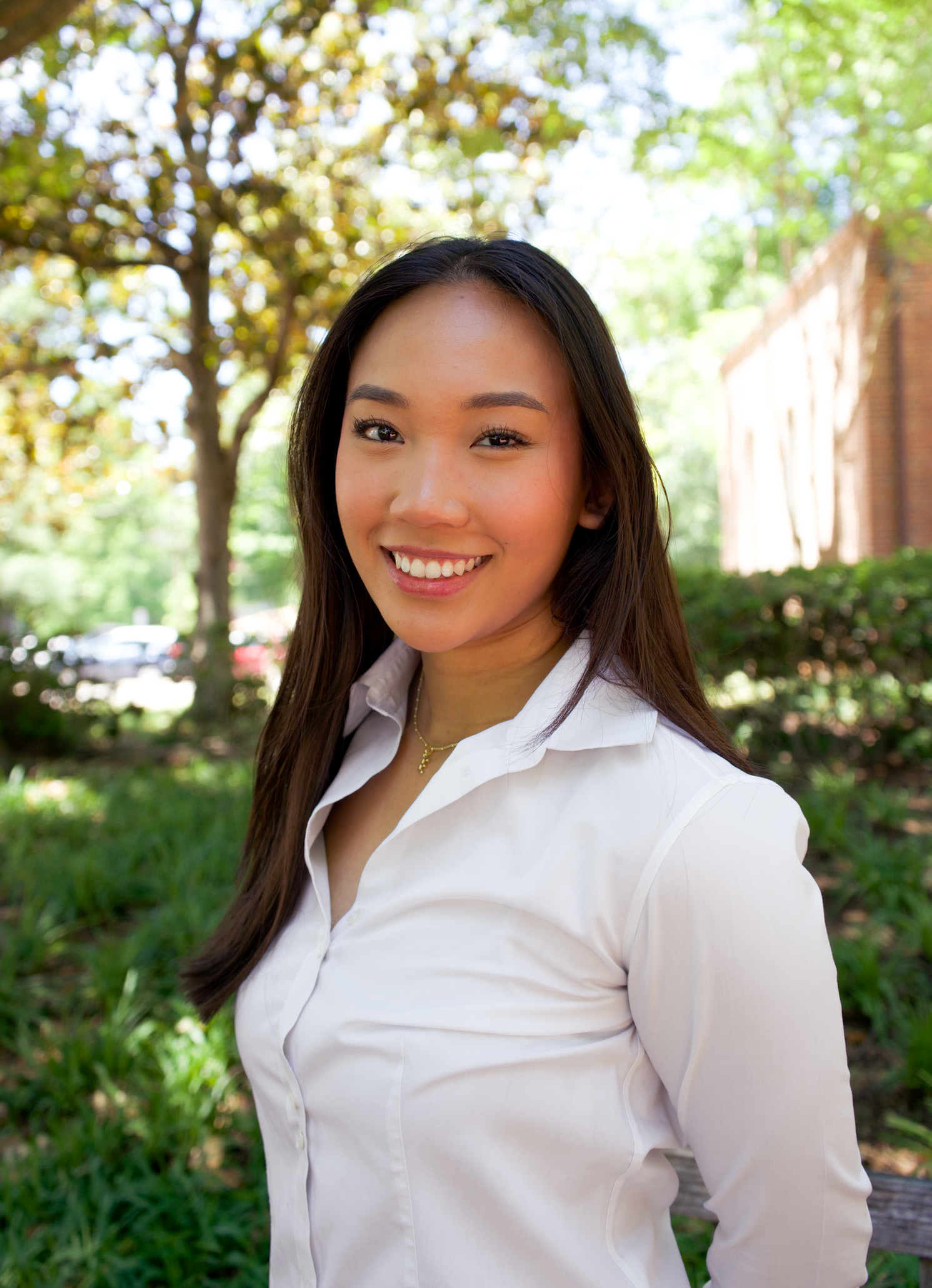 Smiling young woman with long dark hair in white button-up shirt standing outdoors near brick building.