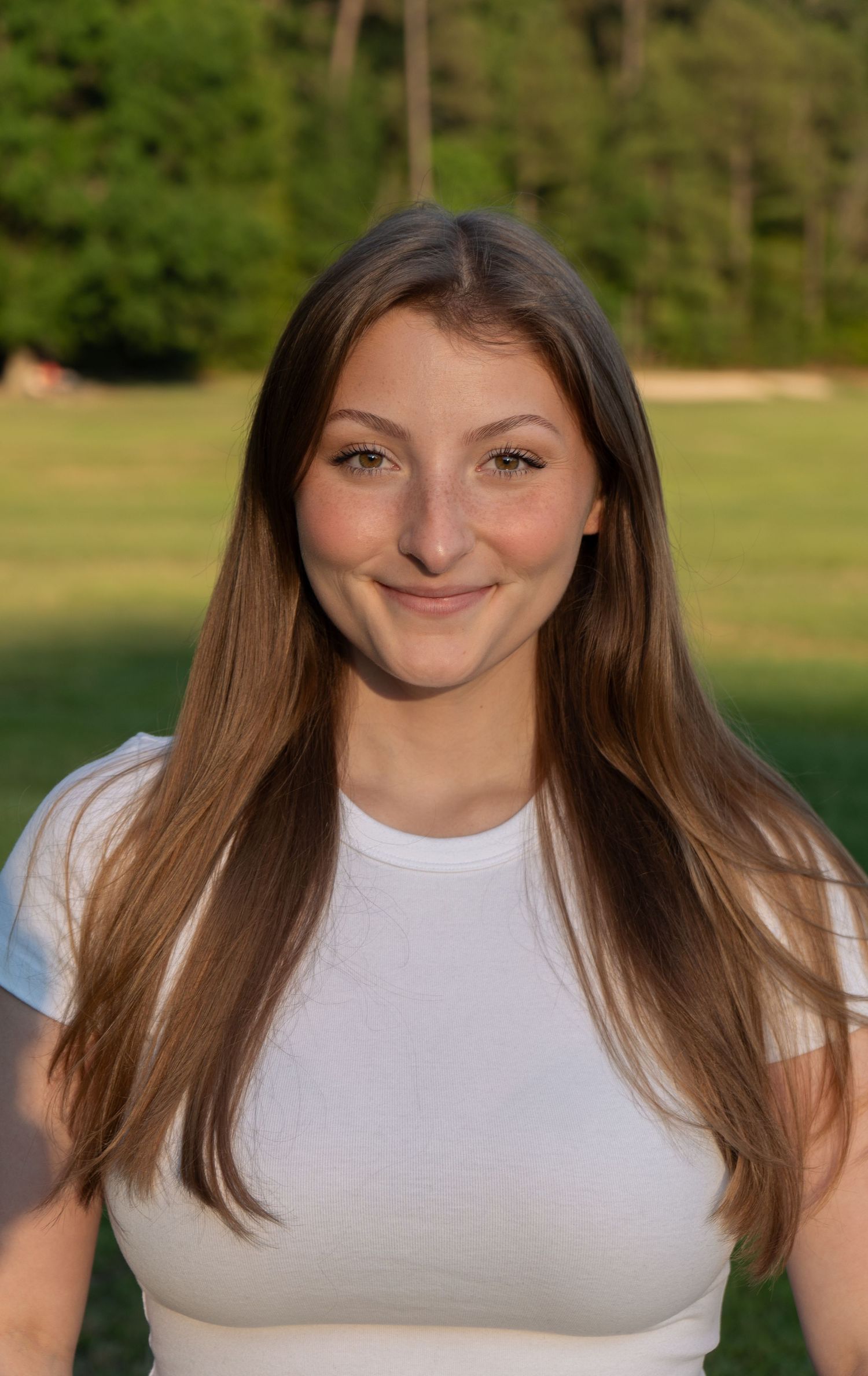 Young woman with long brown hair smiling outdoors in a white top, with green trees in the background.