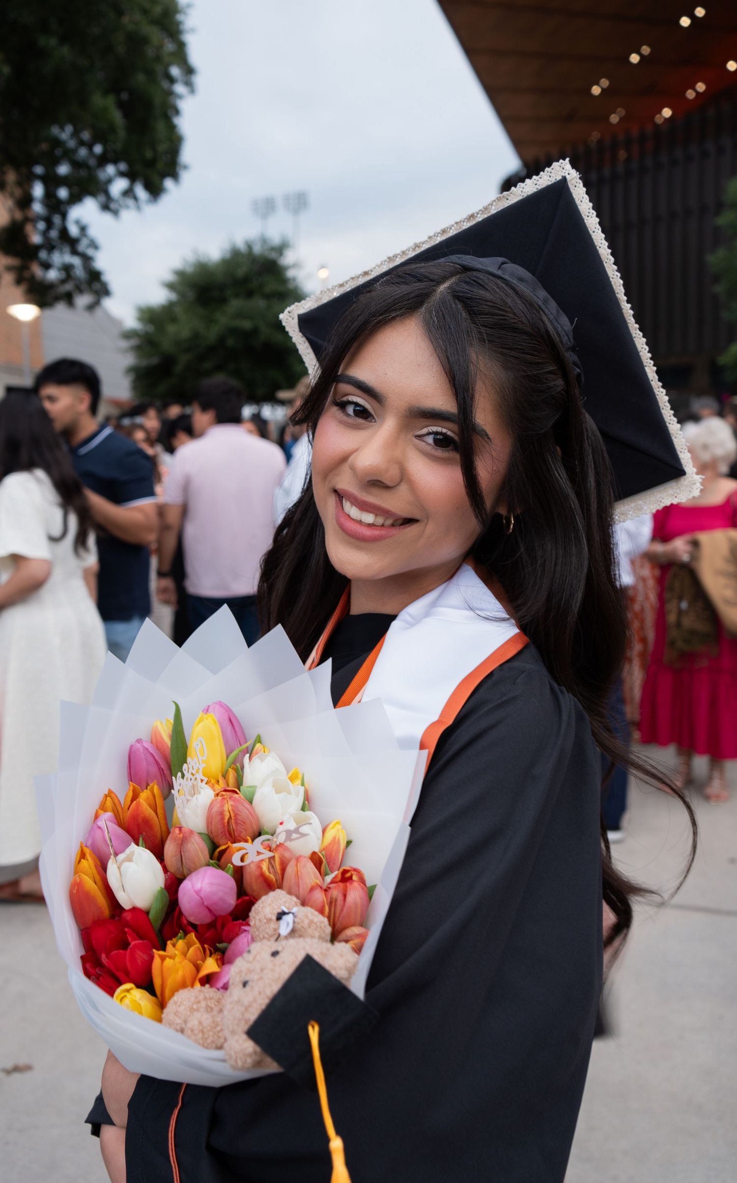 Smiling graduate in cap and gown holds a colorful bouquet of tulips at an outdoor graduation ceremony.