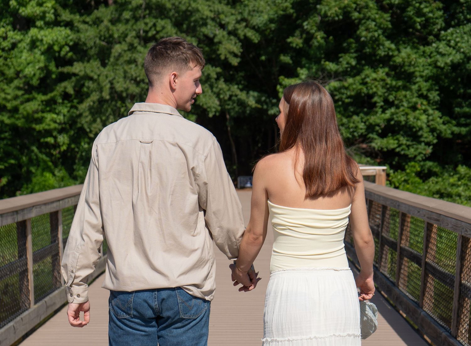 Couple holding hands walking on wooden bridge surrounded by lush green trees on a sunny day.