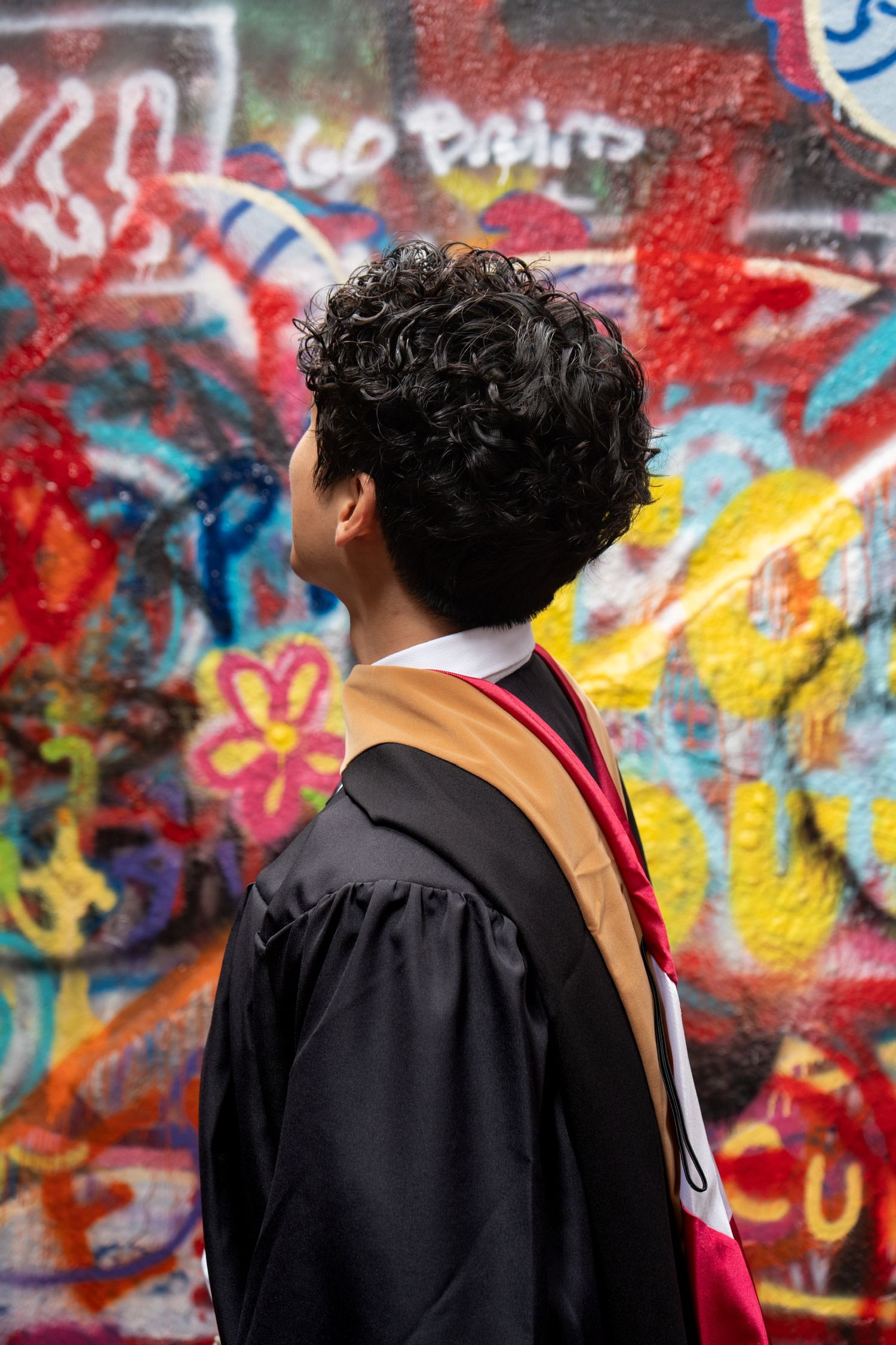 Graduate in black gown with gold stole stands before colorful graffiti wall with Go Spirit text.