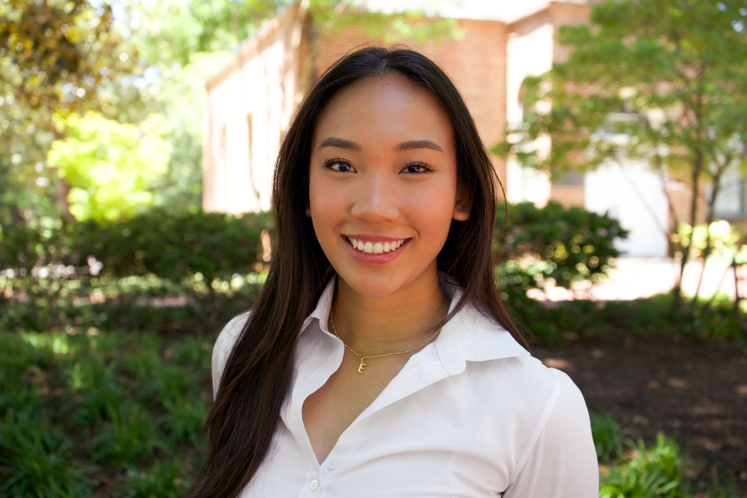 Close-up of a smiling young woman with long dark hair in a white shirt outdoors near brick building.