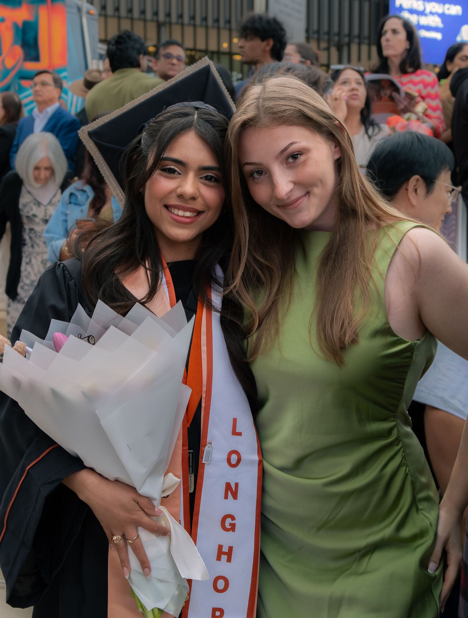 Graduate in cap and gown holding flowers poses with a friend at a university commencement.