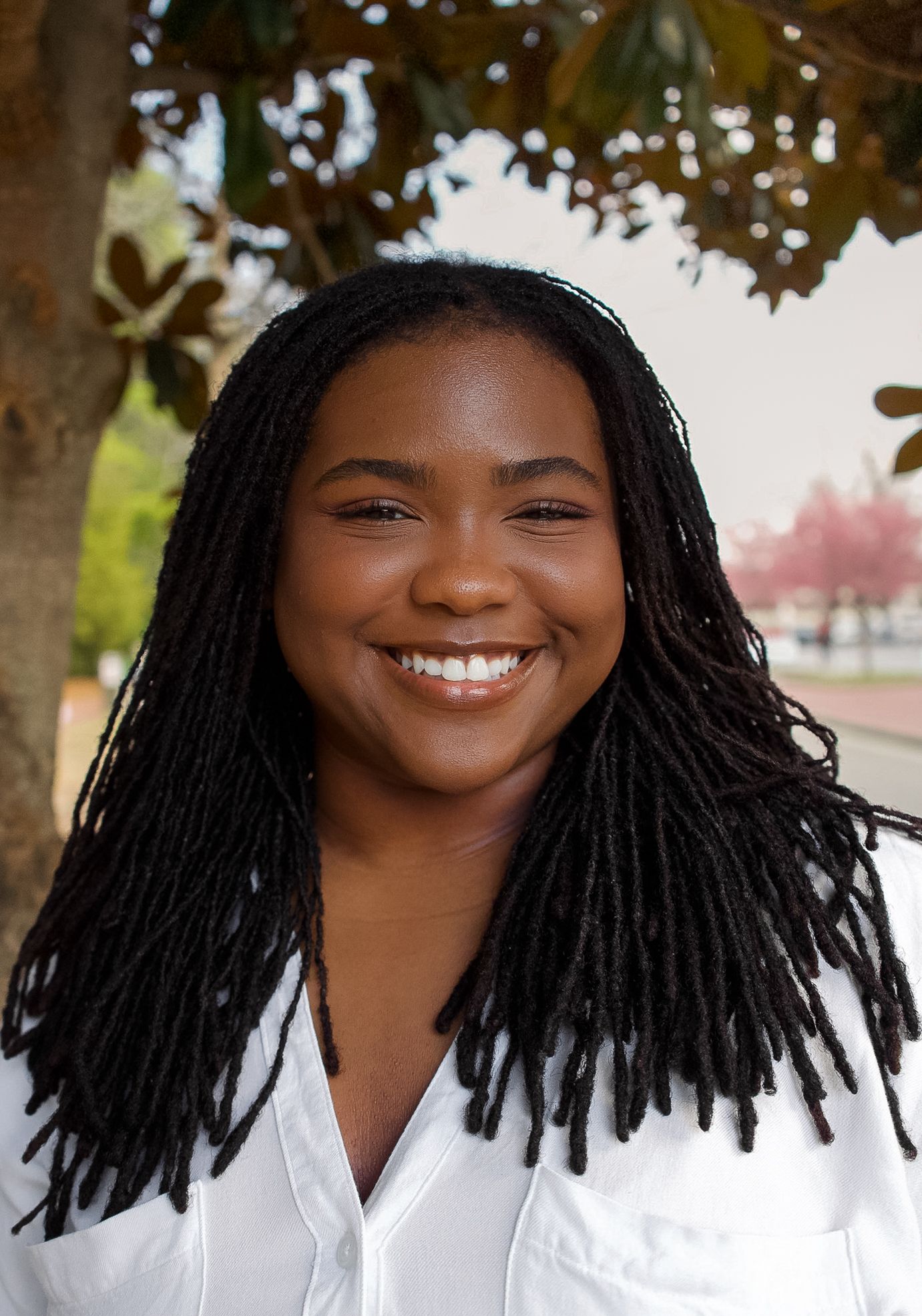 A cheerful young Black woman with long locs smiles warmly while standing under a blooming tree outdoors.