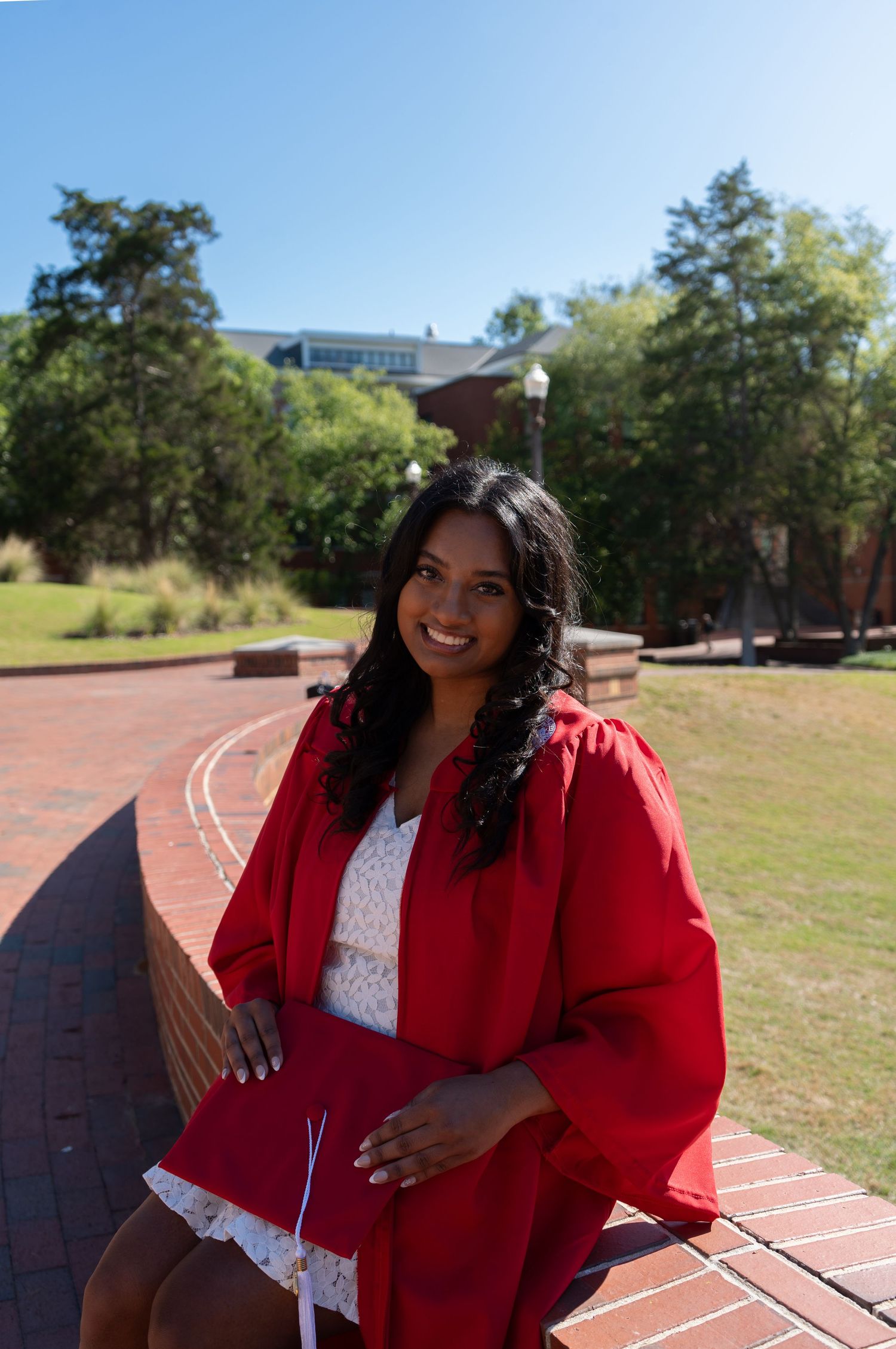 Smiling young woman in red graduation gown sitting on brick steps outdoors on a sunny college campus.