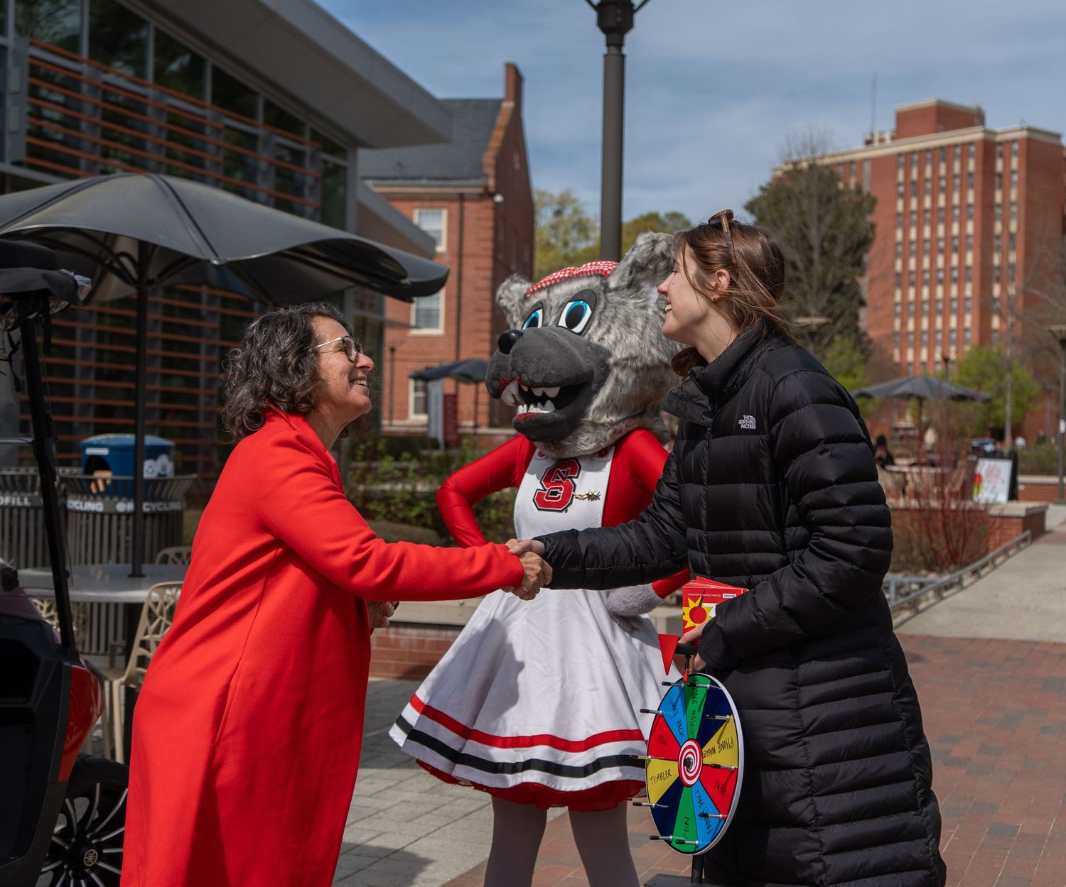 Two women greet a university mascot costume character at an outdoor campus event.