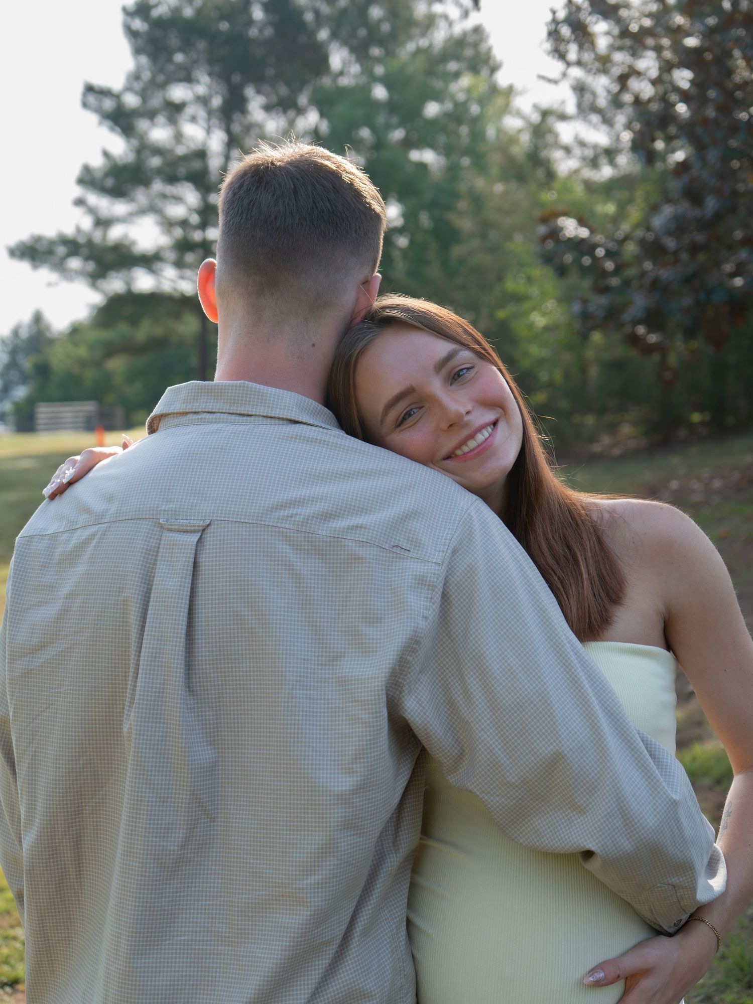 A smiling young woman in a light yellow dress embraces a man in a beige shirt outdoors in a green park setting.