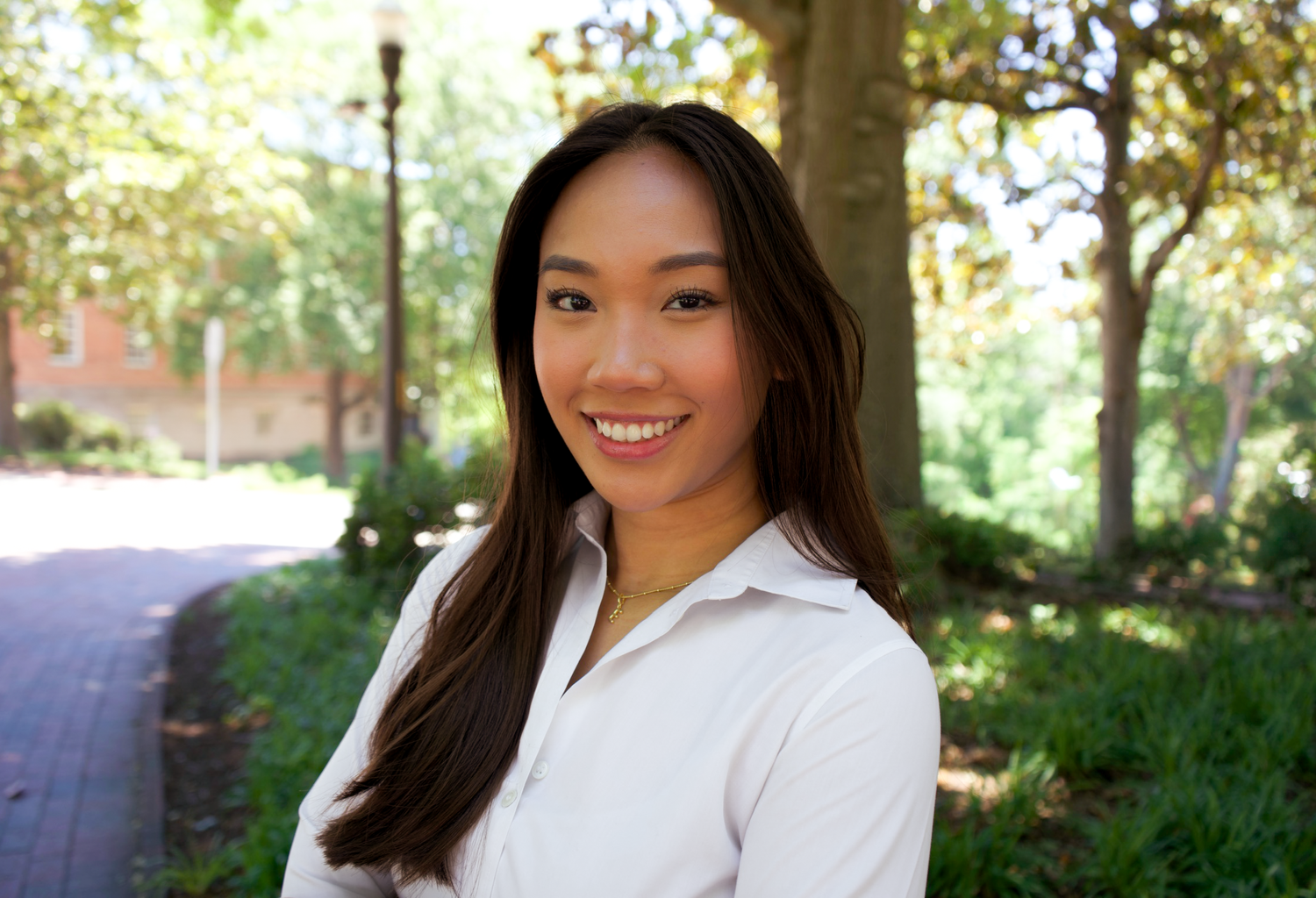 Close-up portrait of a smiling young woman with long dark hair in a white shirt outdoors.