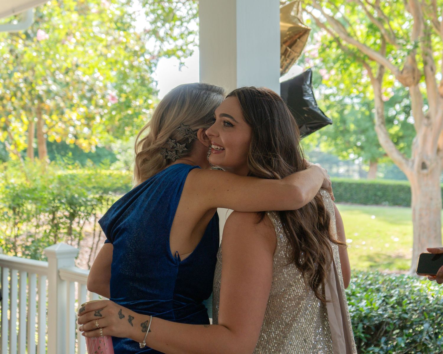 Two women in formal dresses share a warm embrace on a sunlit porch surrounded by greenery.