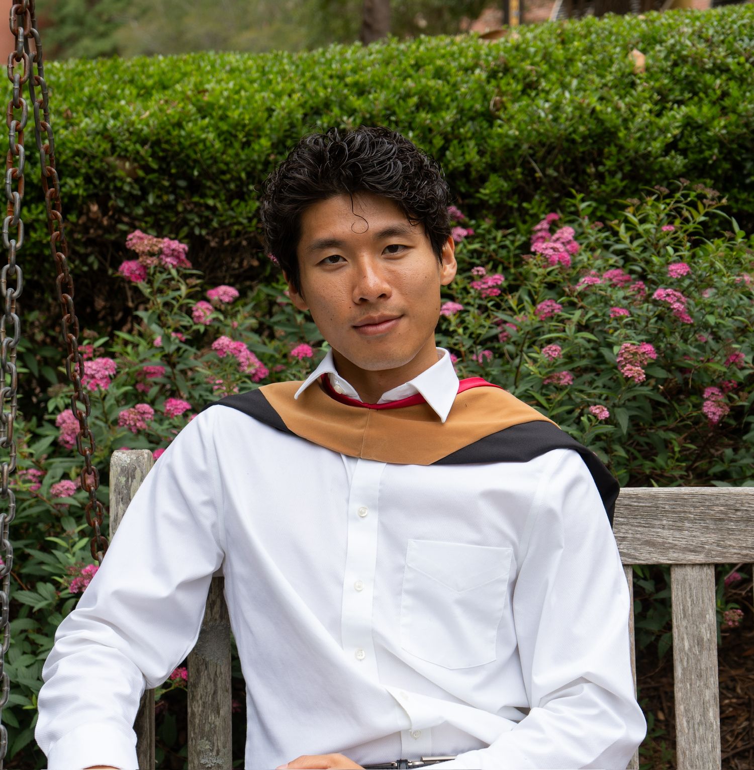 Young man in white shirt with gold and black graduation hood sits relaxed on bench near pink flowers.