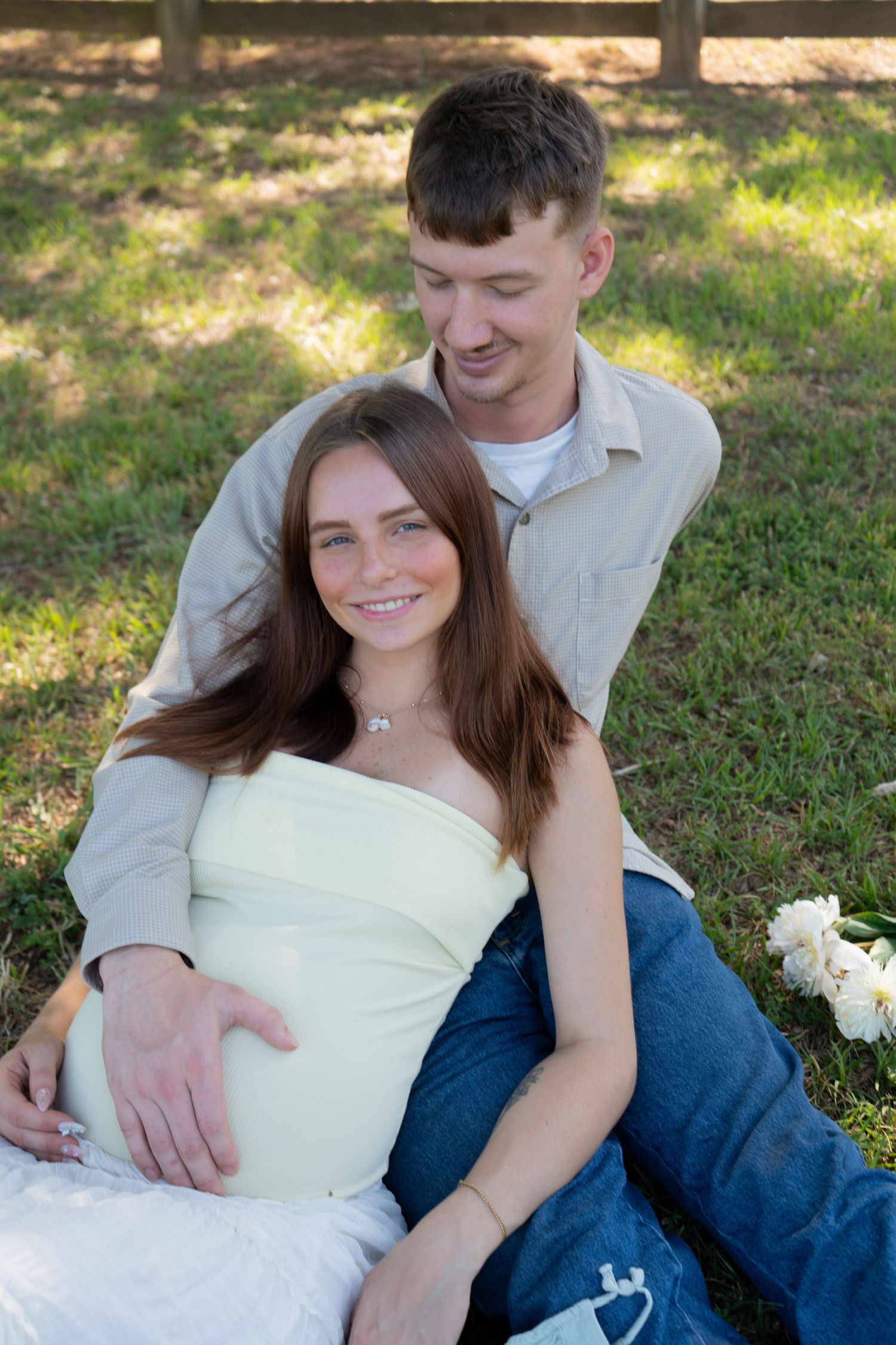 Happy pregnant couple sitting on grass outdoors, woman in white top with partner's hand on baby bump.