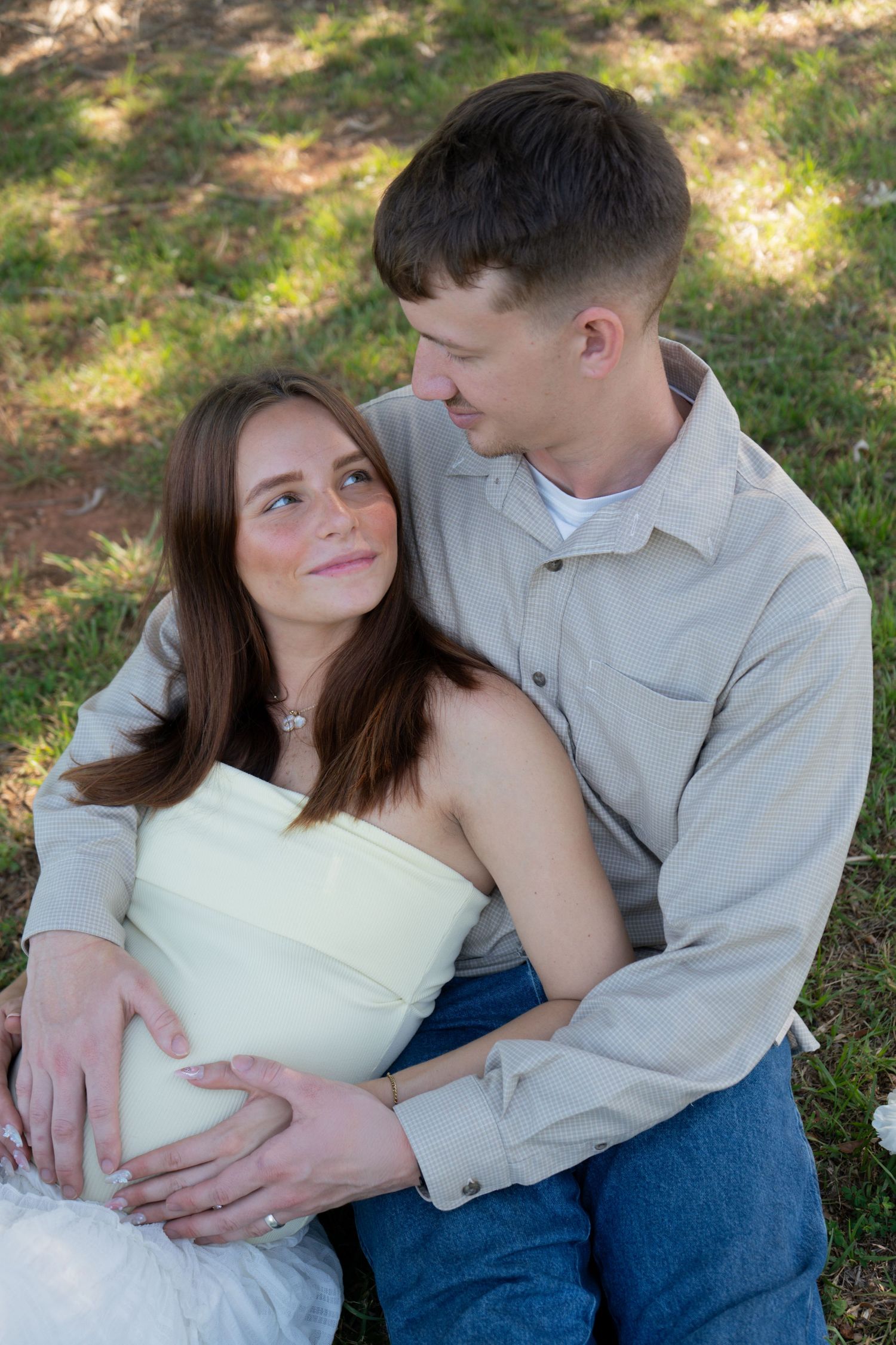 A young couple sits outdoors on grass, the man embracing the pregnant woman from behind in a tender maternity photo.