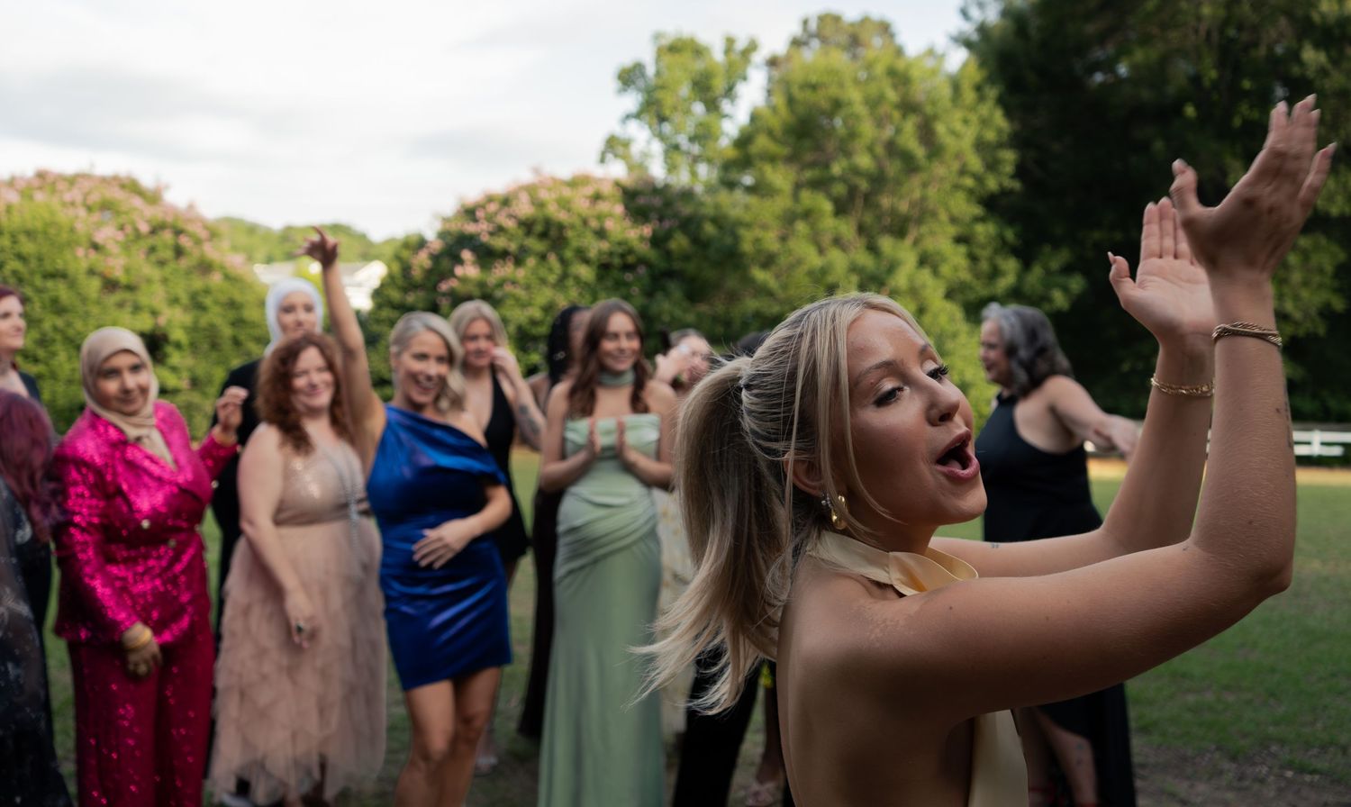 A blonde woman in a gold dress celebrates with raised hands as a group of formally dressed women cheer behind her.