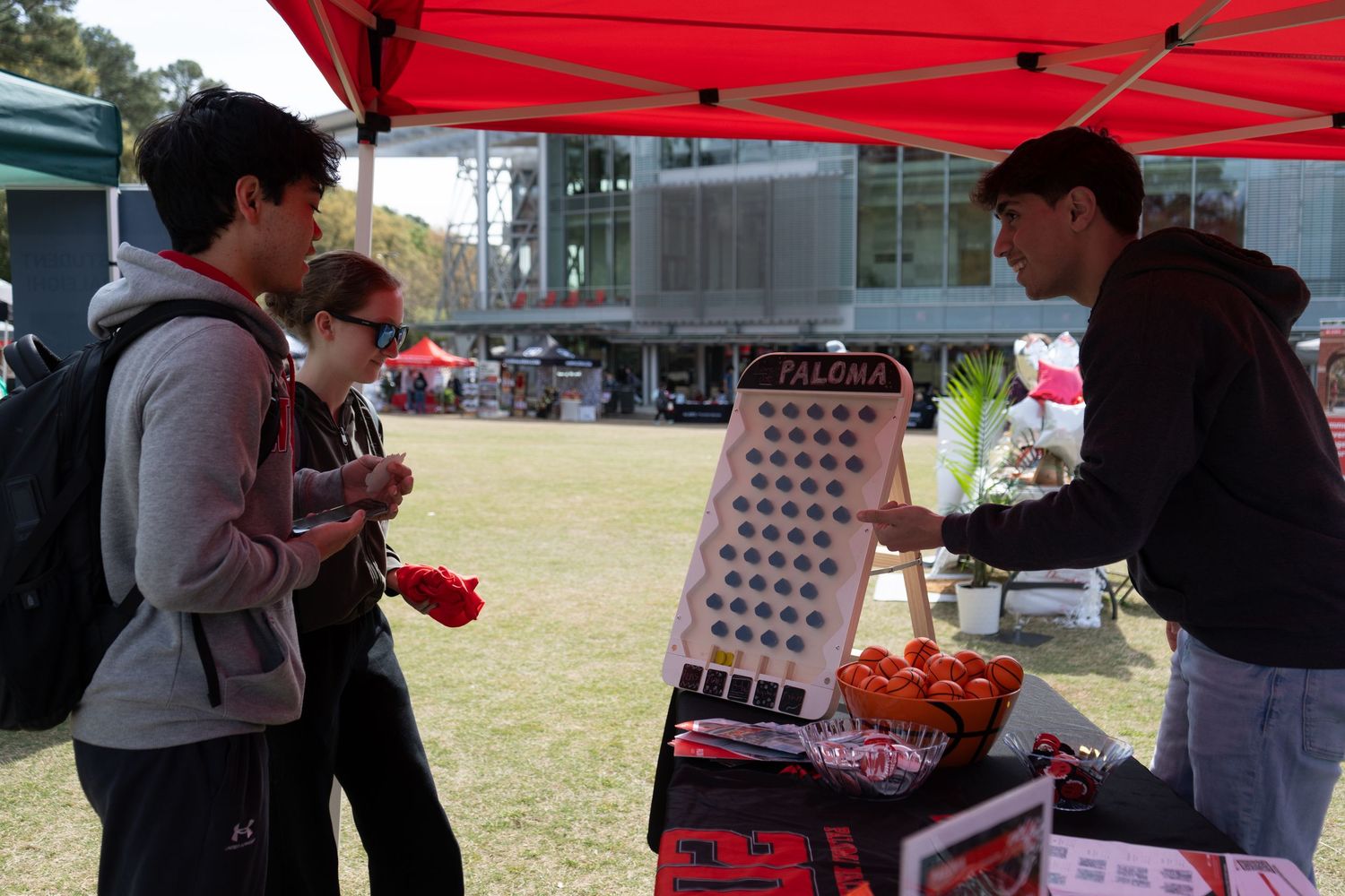Students interact with a vendor at an outdoor campus fair beside a game board display.