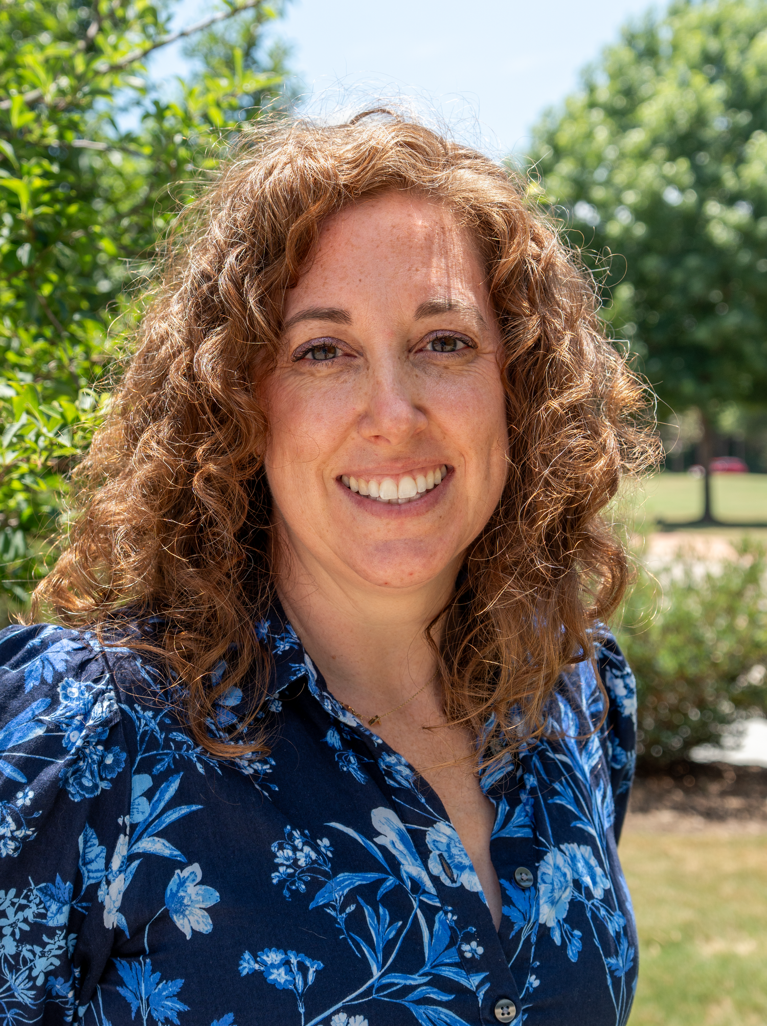 Close-up professional headshot of smiling woman with curly hair in blue floral shirt posing outdoors in sunlight.