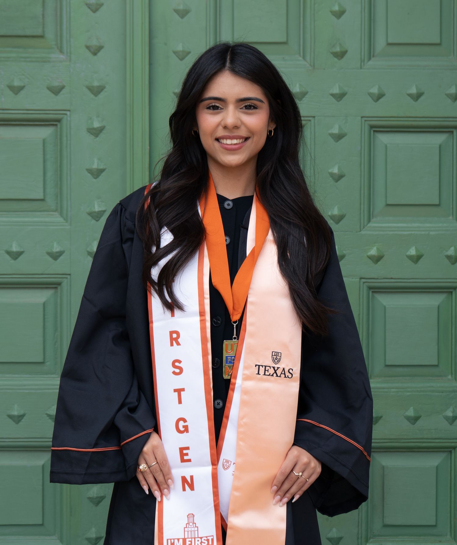 University of Texas graduate smiles in black gown with orange stole and FIRSTGEN sash in front of green doors.