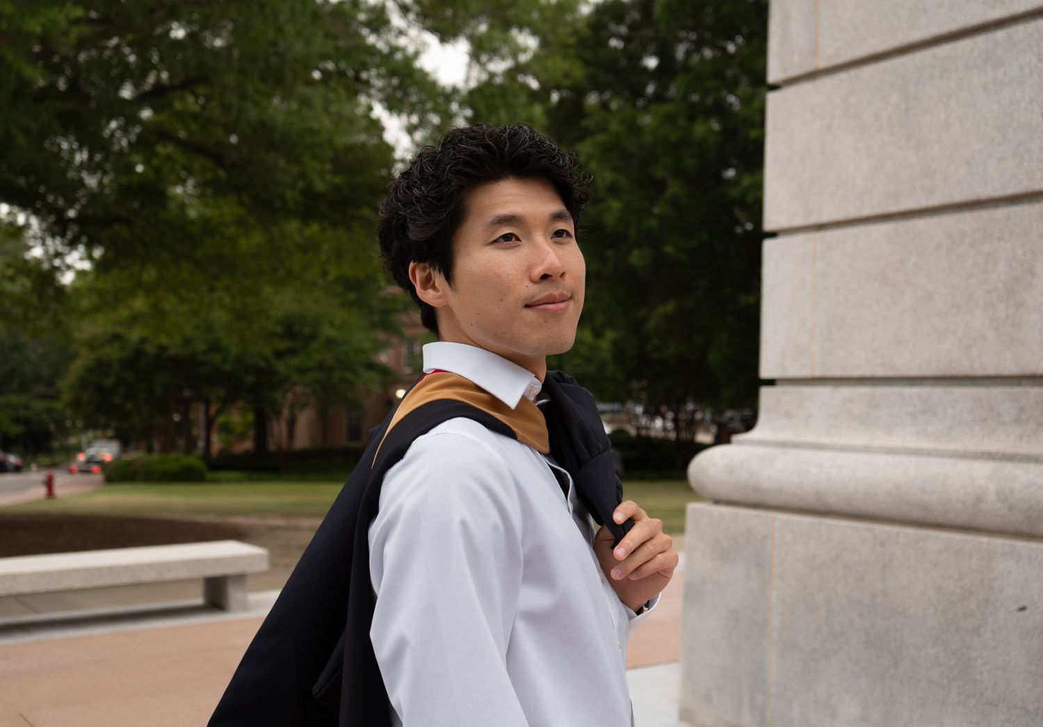 Young Asian man in white shirt with graduation stole smiling while leaning against a column outdoors.