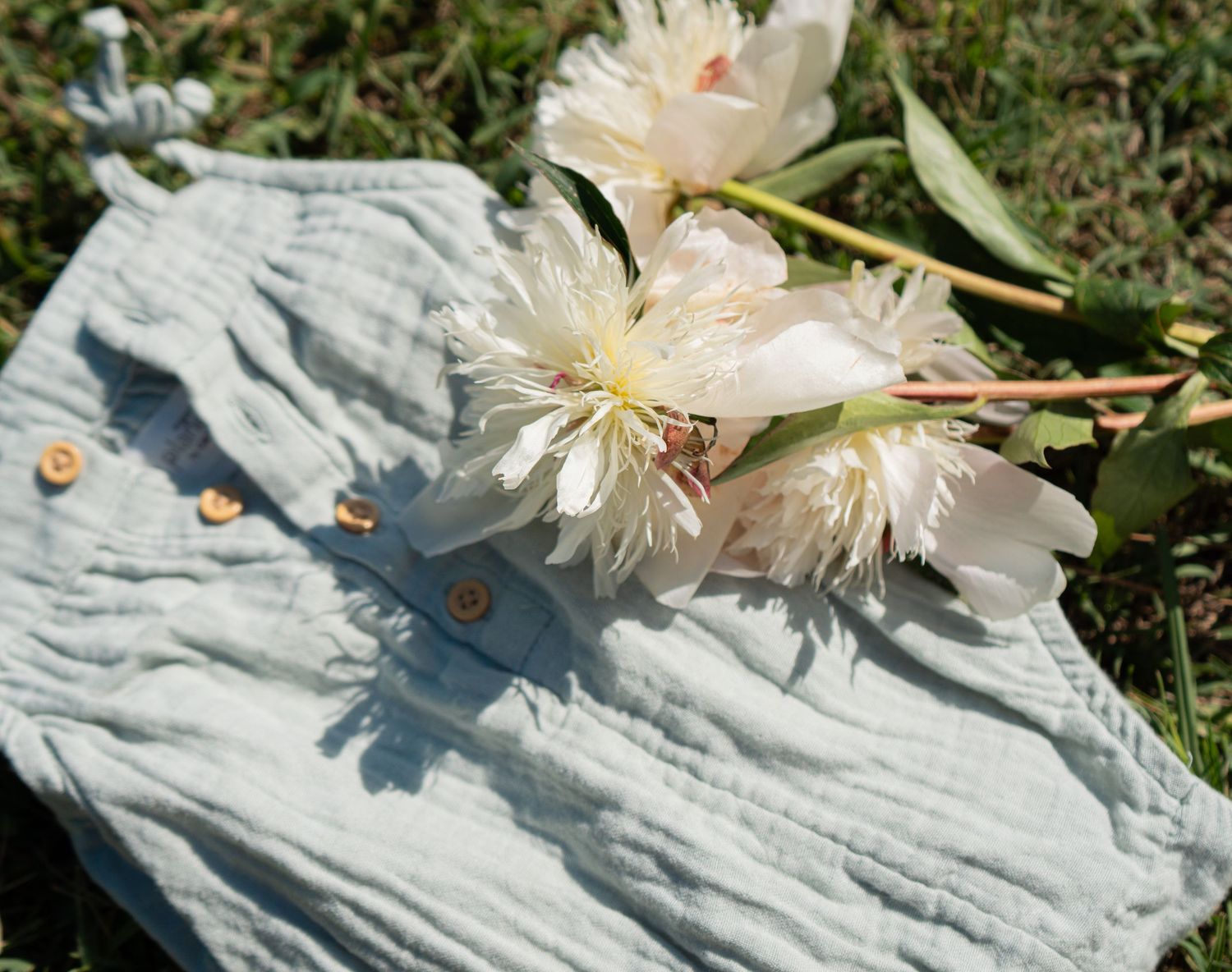 White peony flowers resting on a light blue linen pouch bag outdoors on green grass.