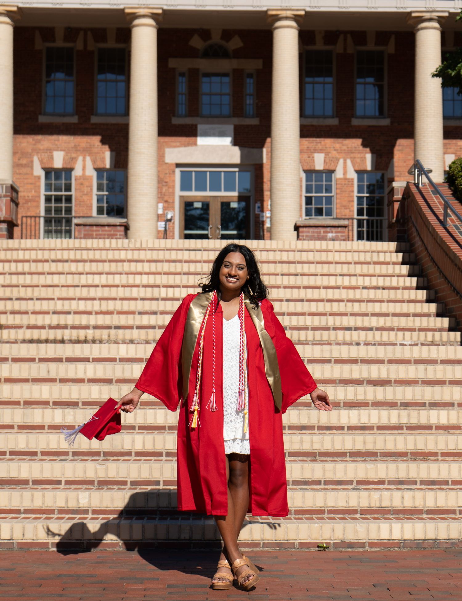 Smiling NC State graduate in red gown with gold stole stands on steps of brick university building.